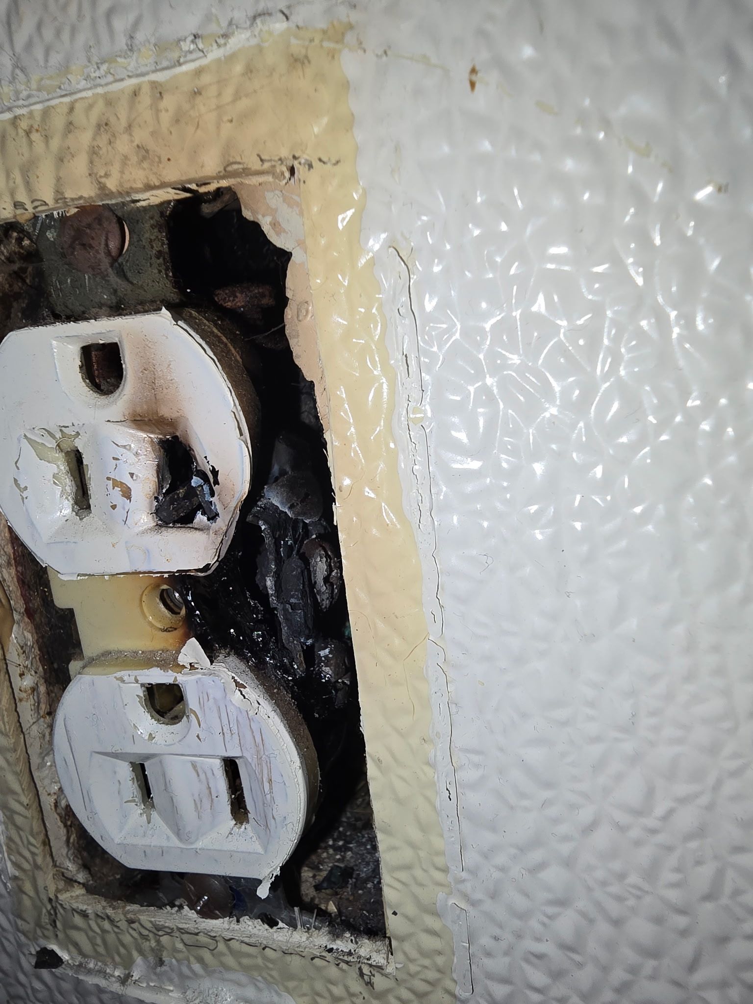 A damaged, charred electrical outlet with melting plastic on a textured, off-white wall.