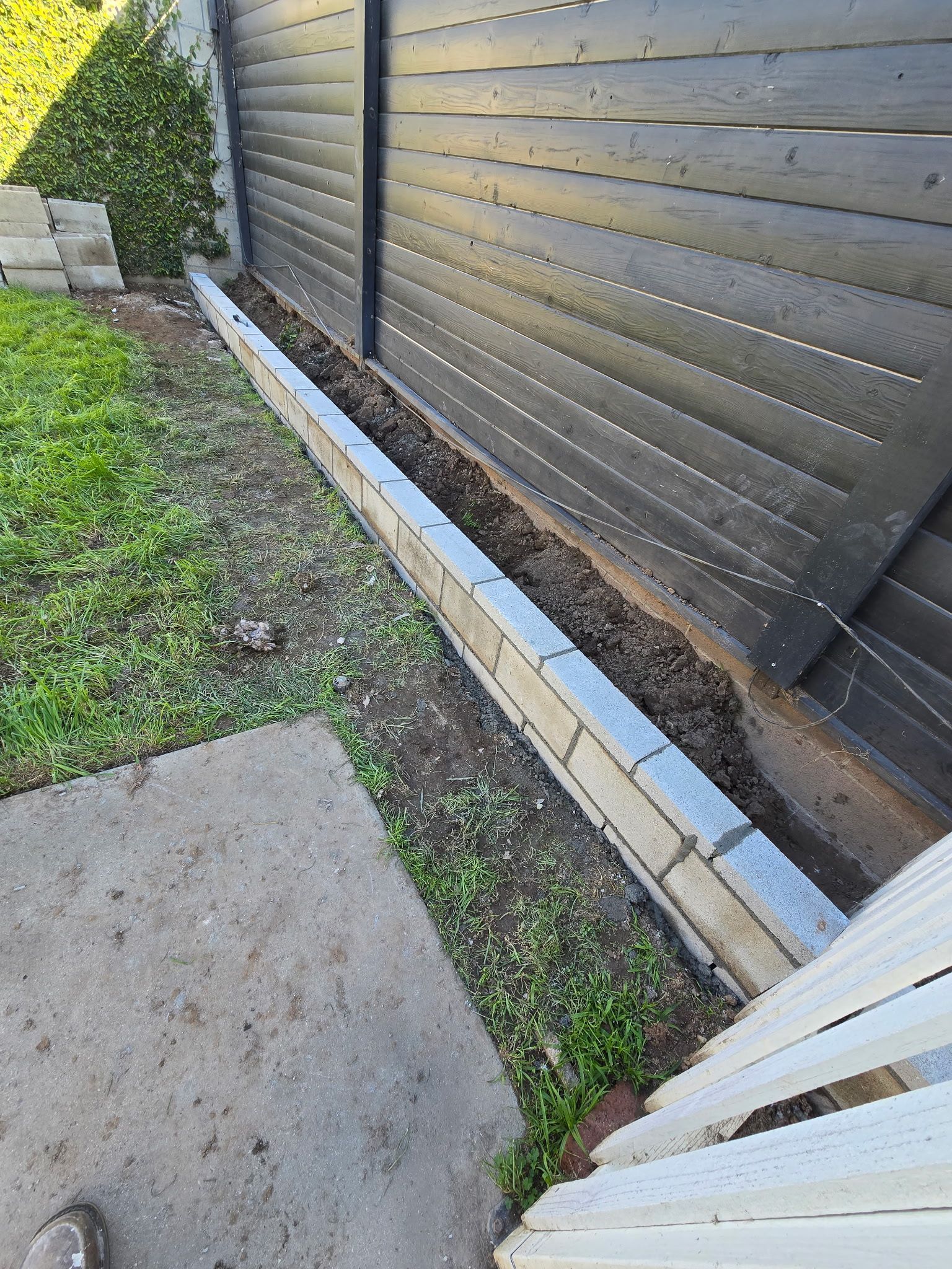 A stone garden border runs along a dark horizontal fence beside a patch of grass and a concrete walkway.