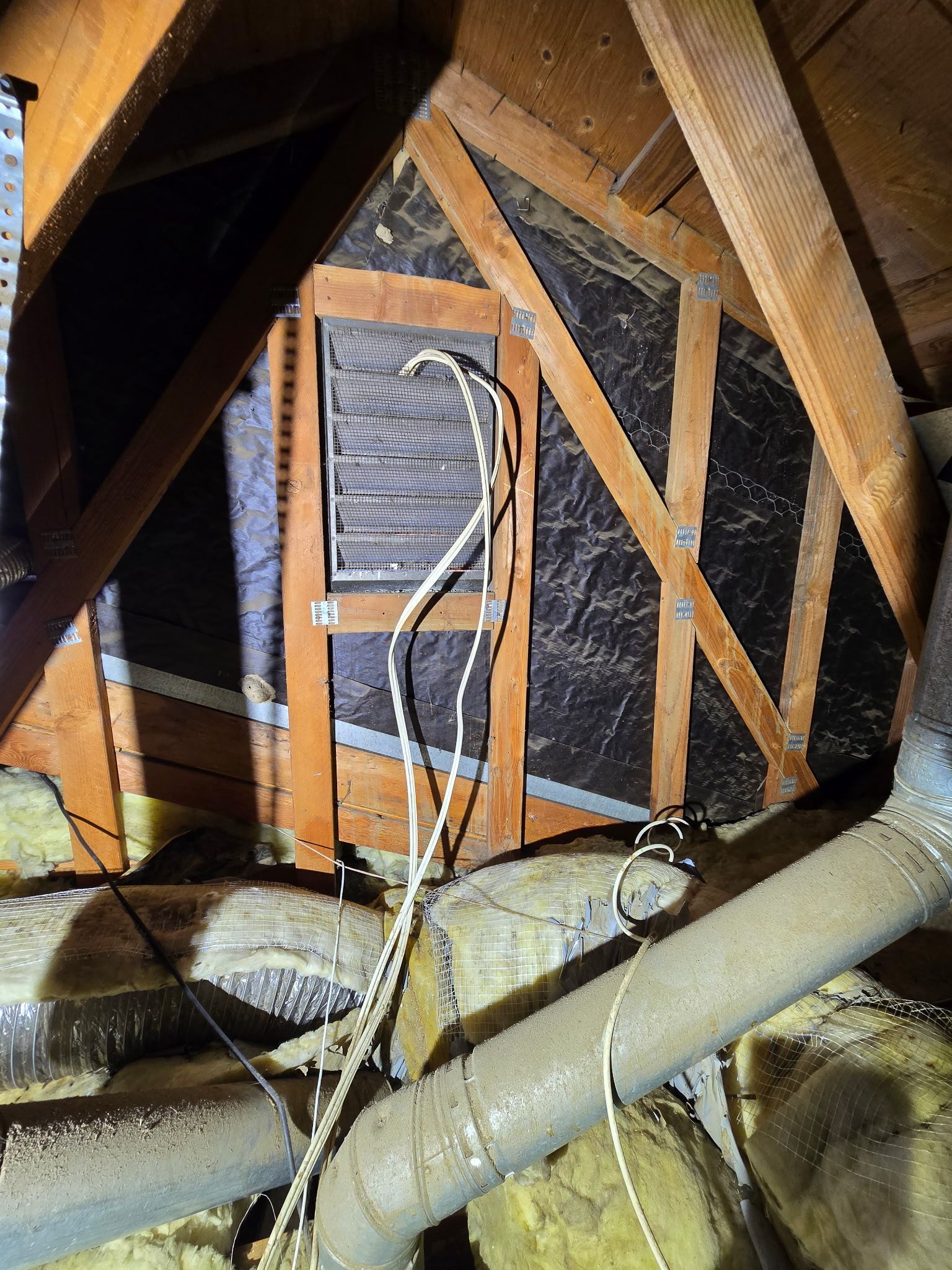 View of an attic space showing wooden rafters, a vent in the gable wall, insulation, and electrical wiring.