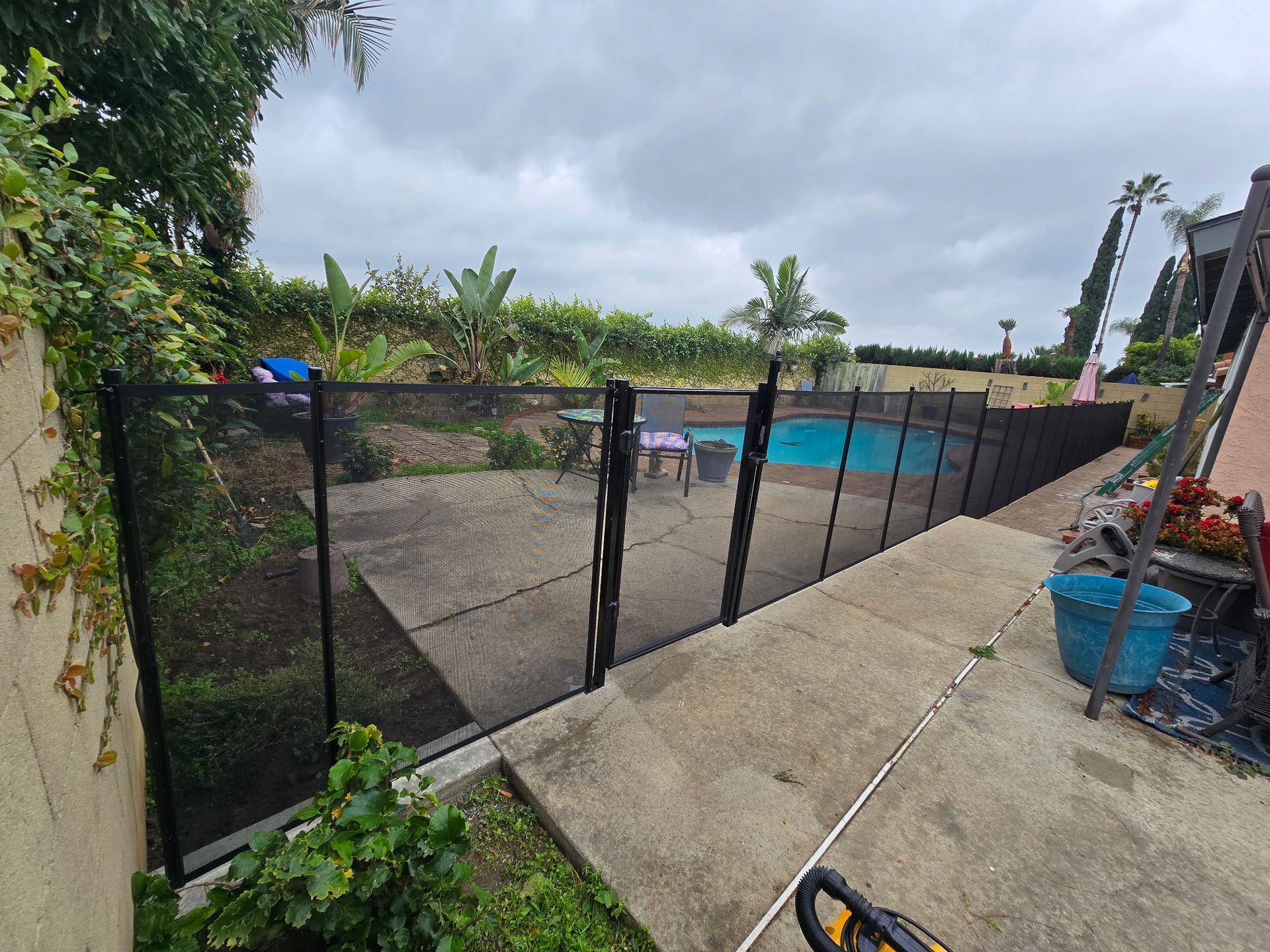 A mesh safety fence surrounds a backyard swimming pool, installed on a concrete patio under a cloudy sky.
