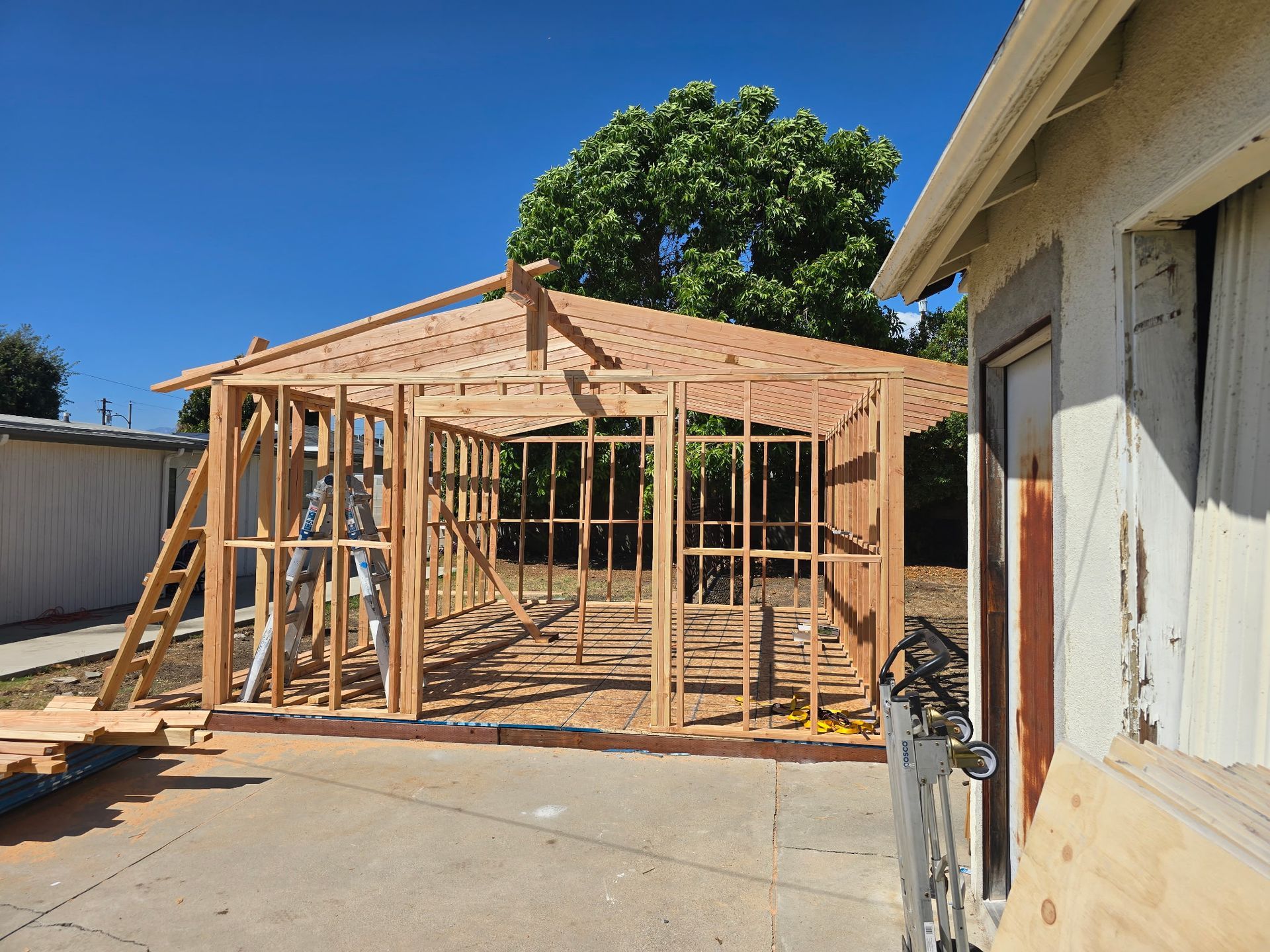 Wood-framed garage/shed under construction in a sunny backyard beside a white building