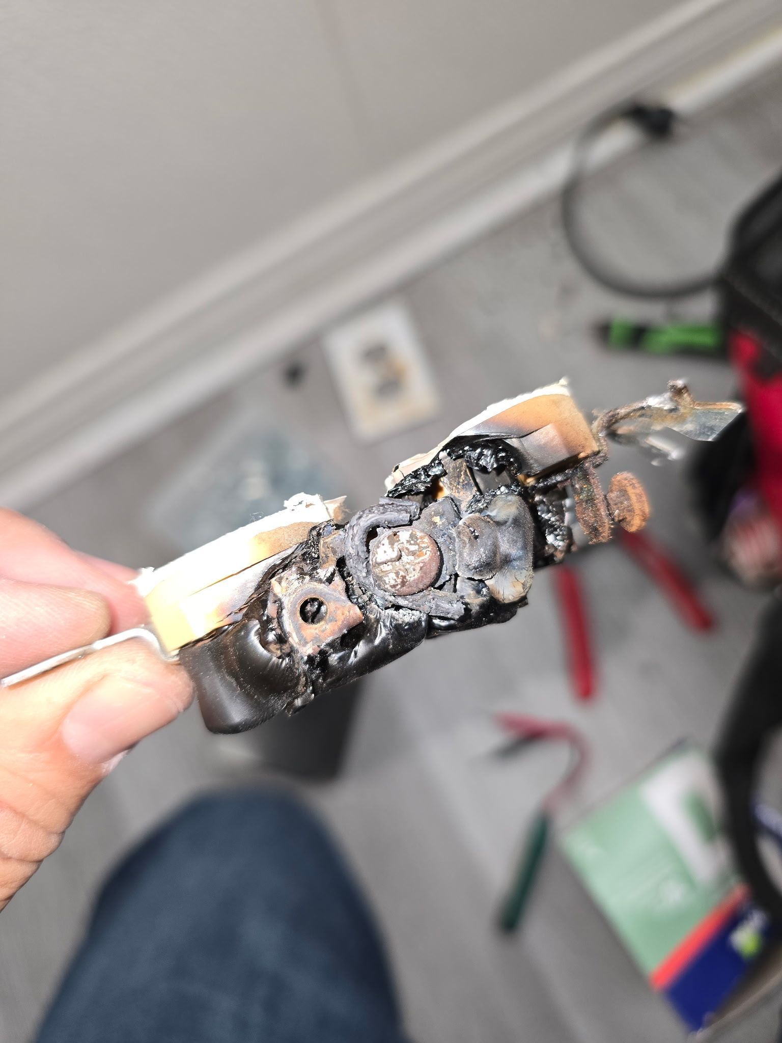 A person holds a severely melted, charred electrical terminal block with damaged wire connections and burnt plastic casing.