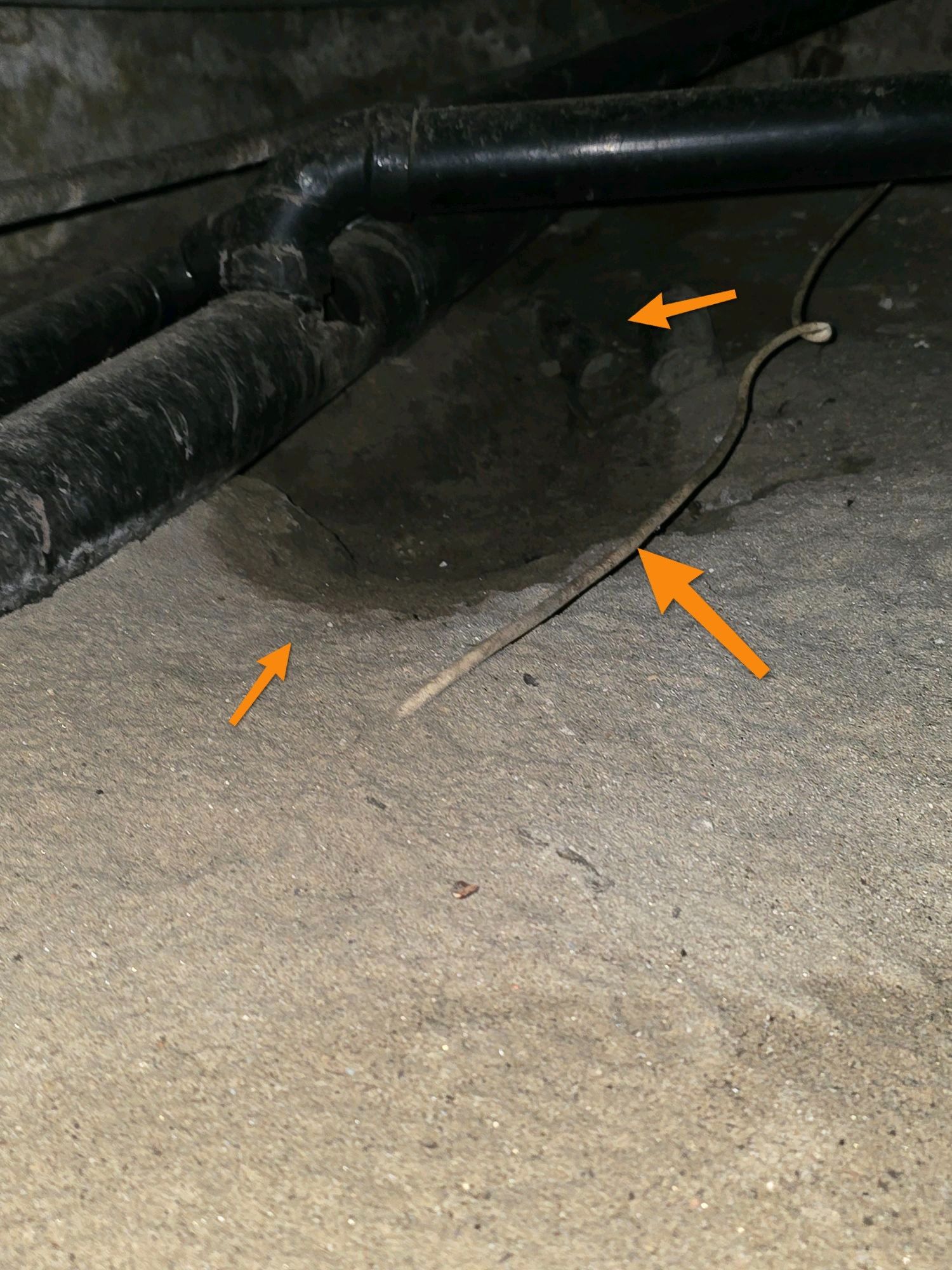 A hole in a crawl space dirt floor beneath plumbing pipes, marked by three orange arrows.