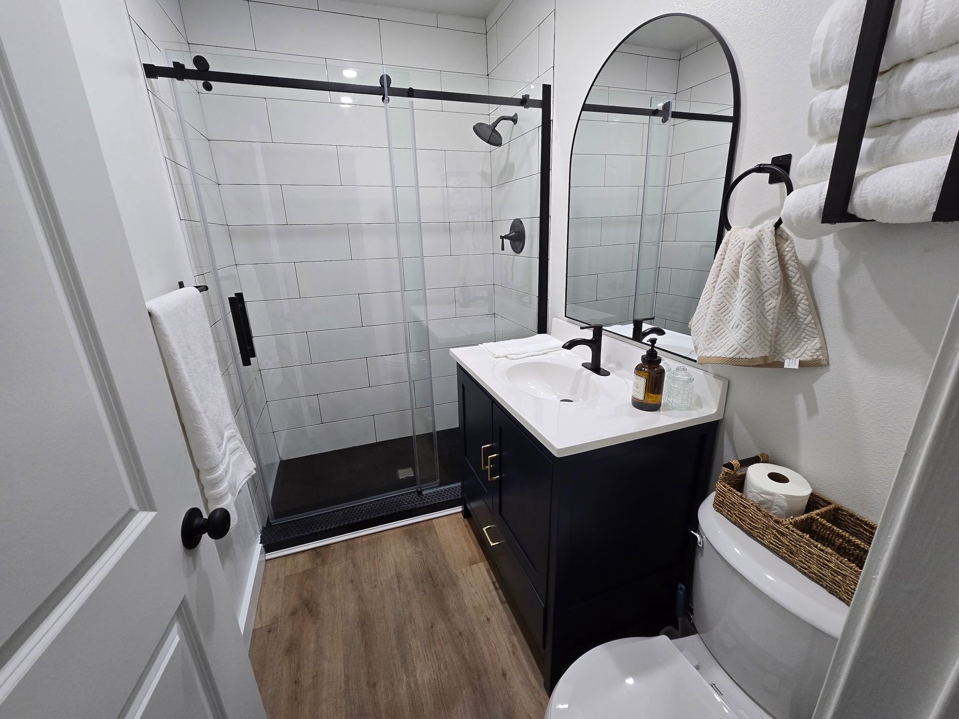 Modern bathroom with a black vanity, white subway tile shower, arched mirror, and wood-look flooring.