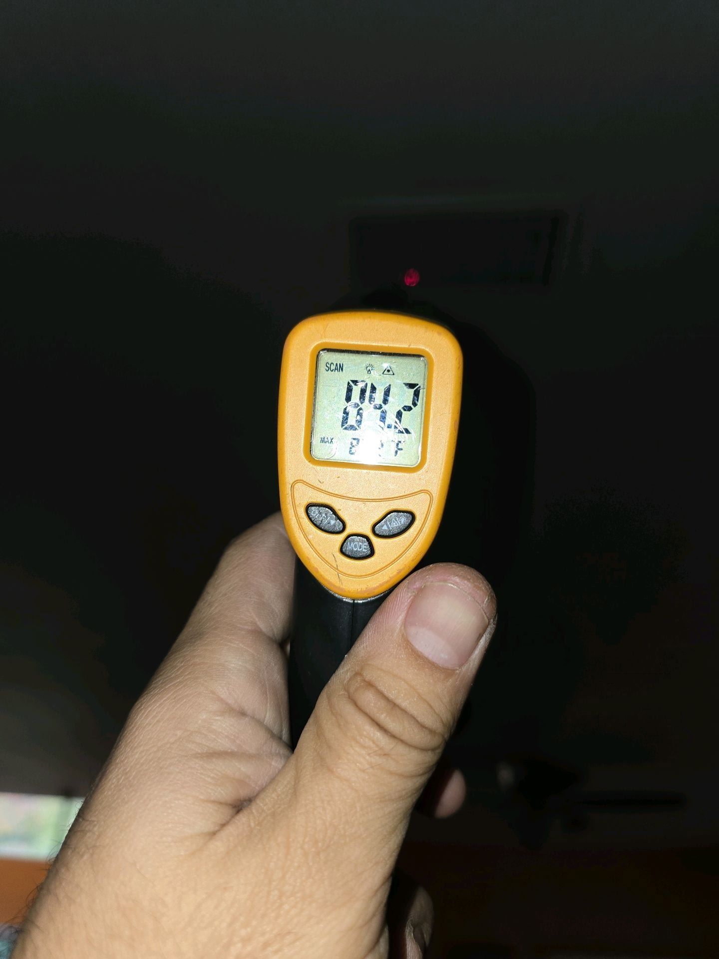 A hand holds a yellow infrared thermometer displaying 84.2 degrees, aimed at an air conditioner unit on a wall.