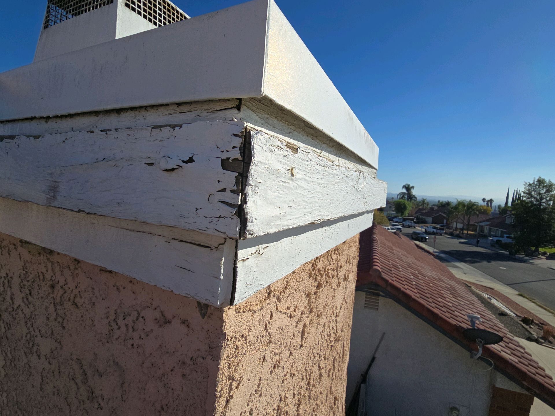 A close-up of a damaged chimney top with peeling white paint and separated wooden trim on a sunny day.