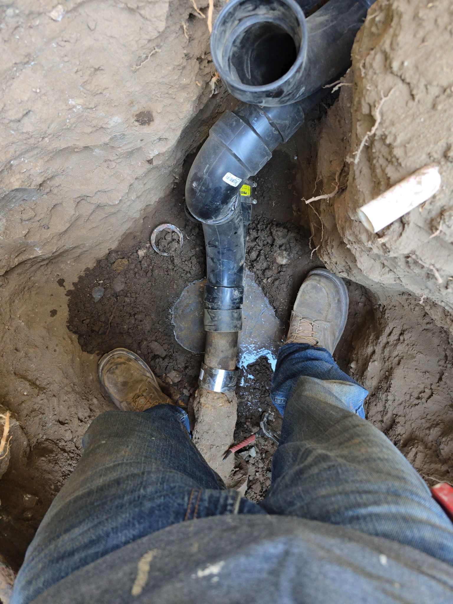 A person in work boots stands in a trench next to a newly connected black plastic sewer pipe fitting.