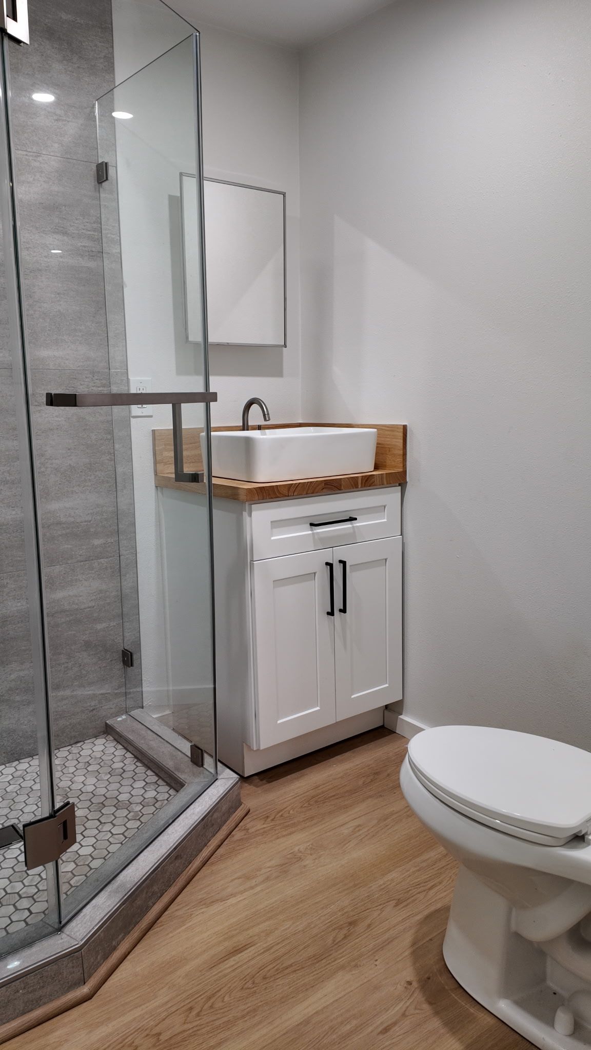 A small modern bathroom featuring a white vanity with a vessel sink, a corner glass shower, and a white toilet.