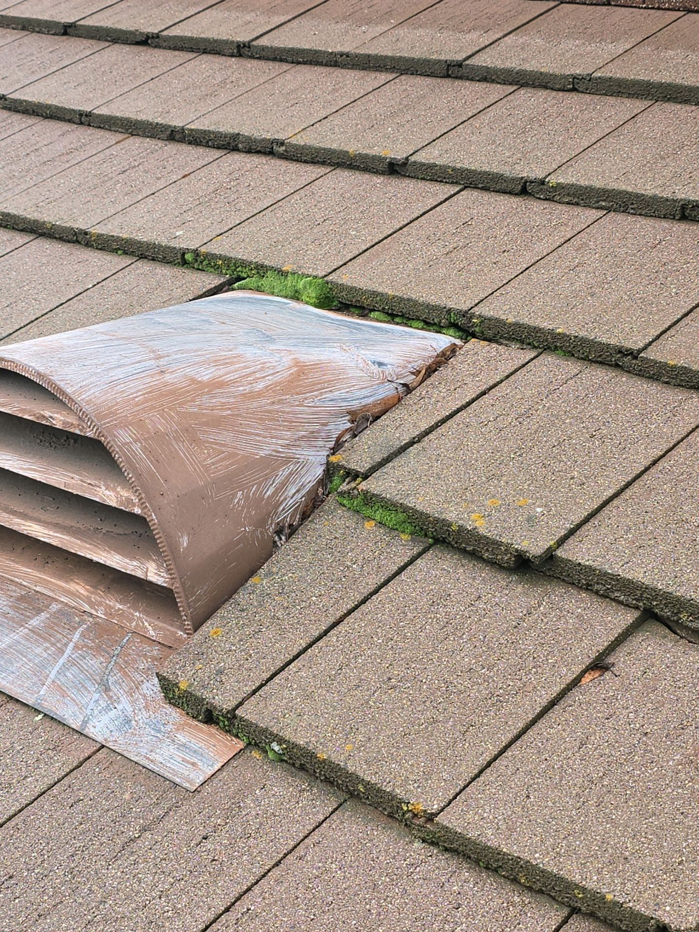A roof vent with weathered brown louvers installed on a brown tiled roof, with visible moss growth in the gaps.