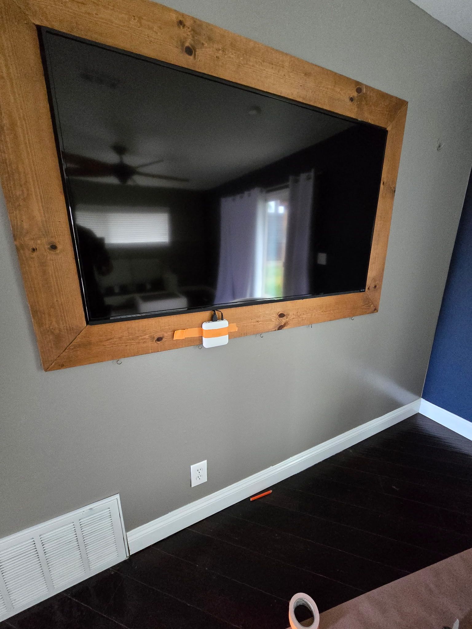 A wall-mounted flat-screen TV framed in rustic wood, with tape visible on the bottom edge, against a grey wall.