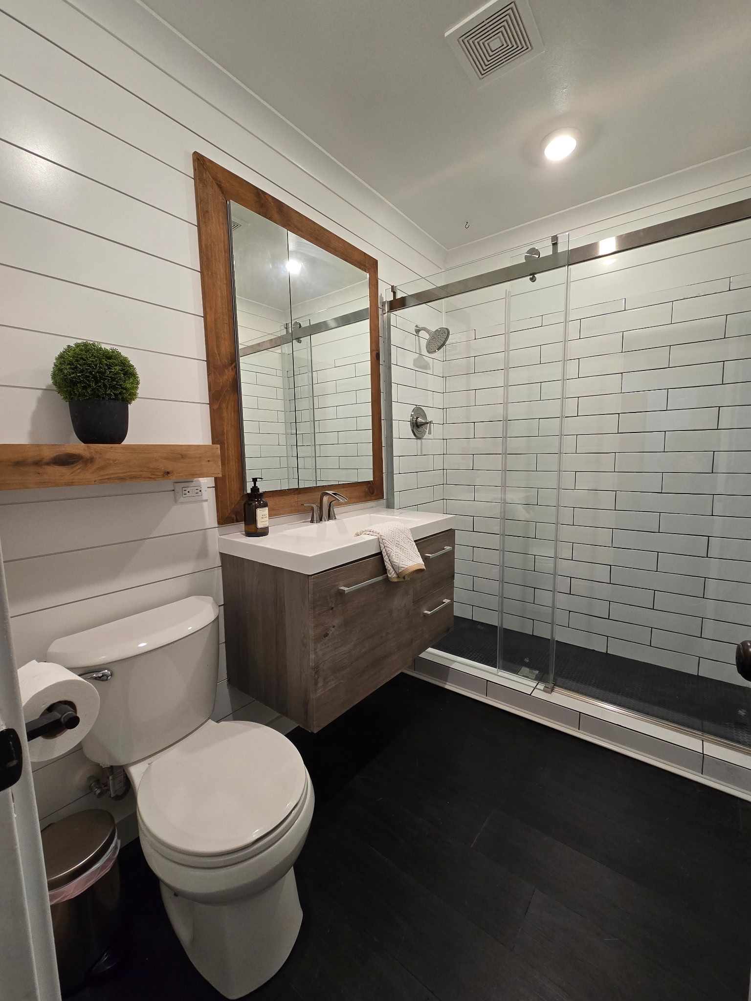 A modern bathroom with white shiplap walls, a floating wood vanity, a rustic mirror, and a glass-enclosed shower.