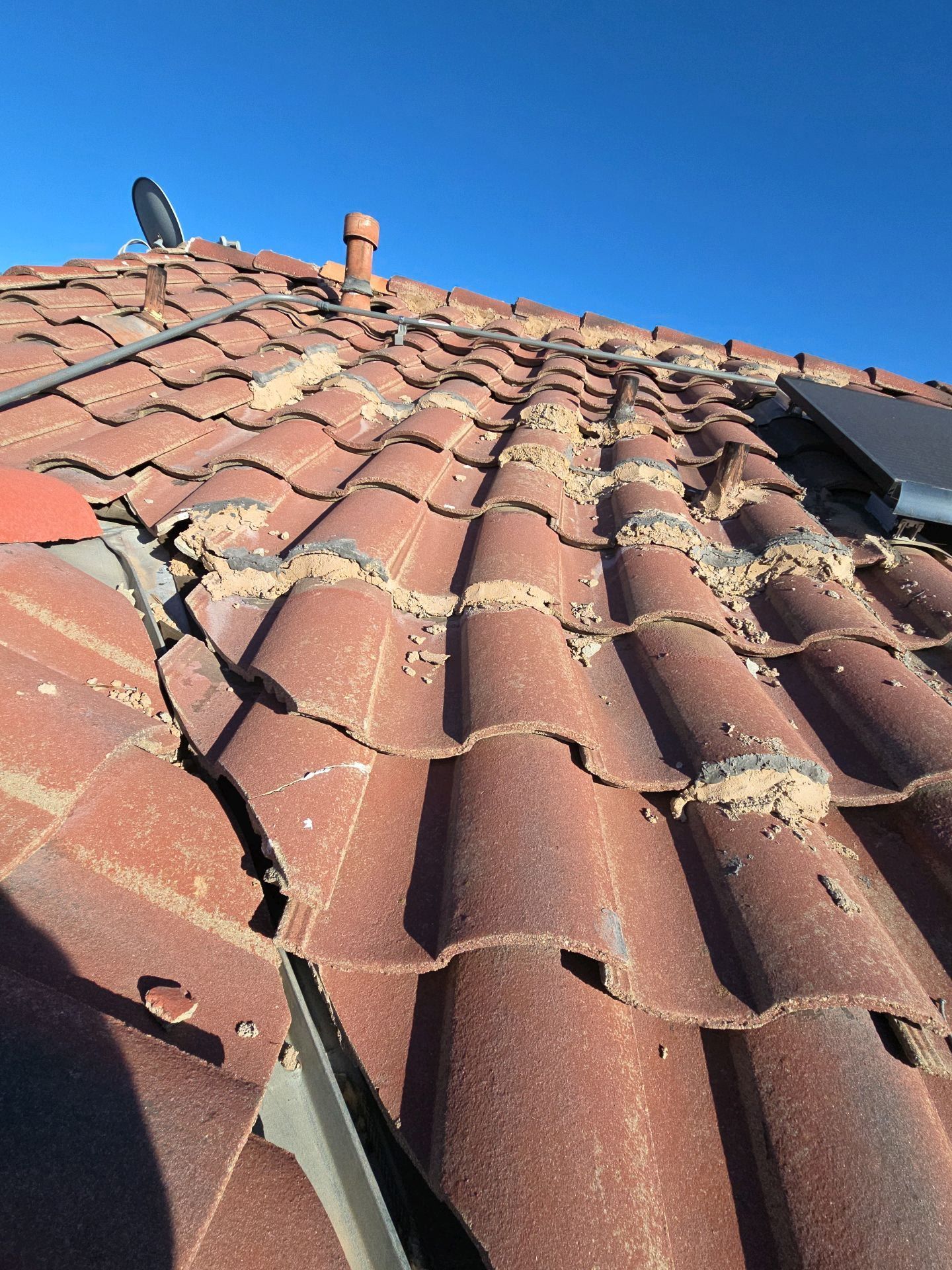 A tiled residential roof under a clear blue sky, showing cracked and weathered tiles with crumbling mortar bedding.