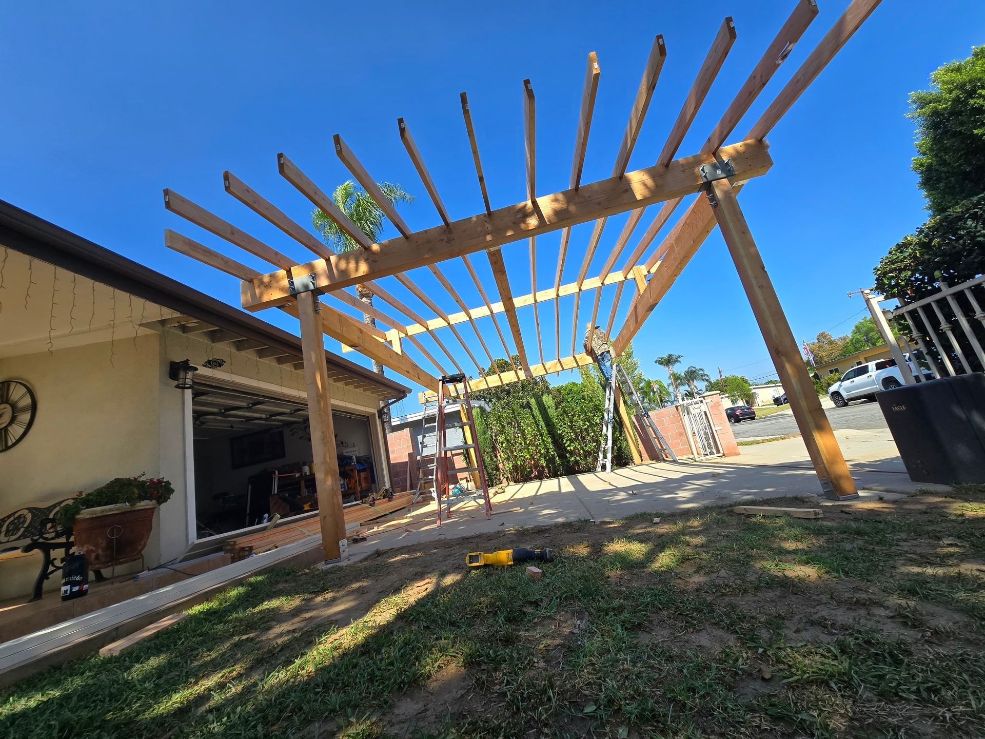 Wooden carport frame in a sunny backyard beside a house, with grass and patio furniture.
