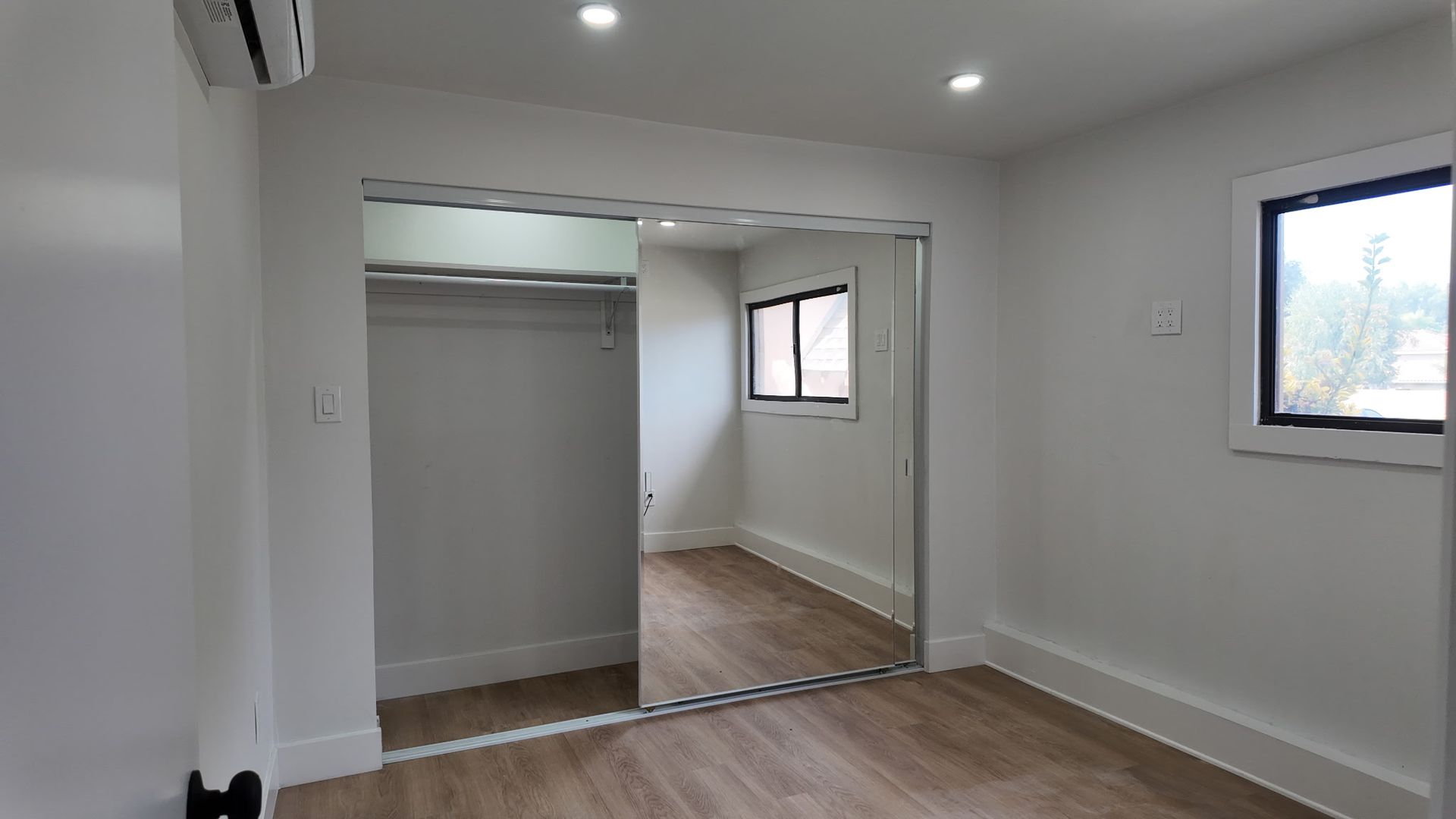 A bright, minimalist bedroom with white walls, light wood-look flooring, a mirror-sliding closet, and a small window.