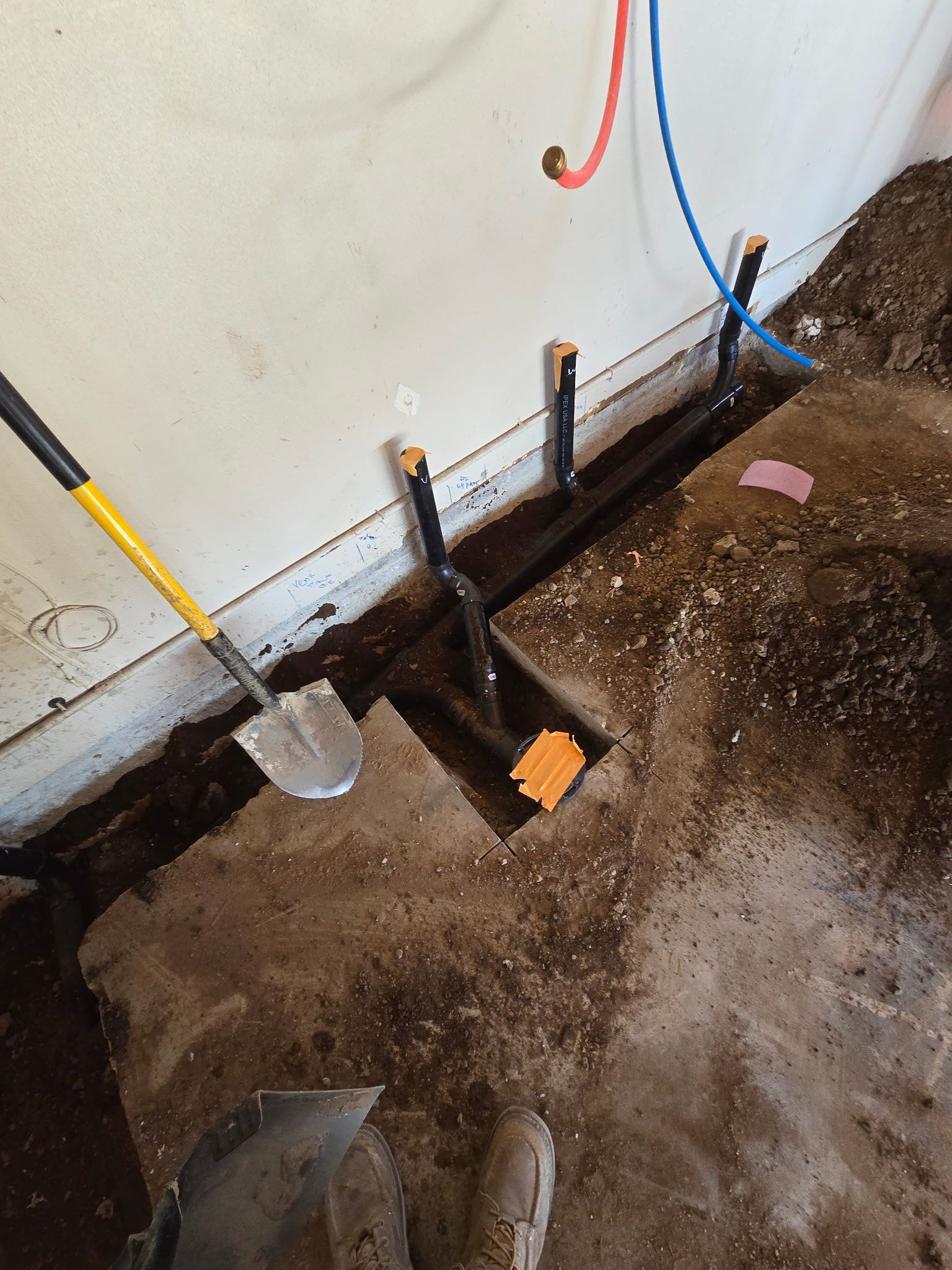Plumbing pipes installed in a trench cut through a concrete floor, with a shovel nearby against a white wall.