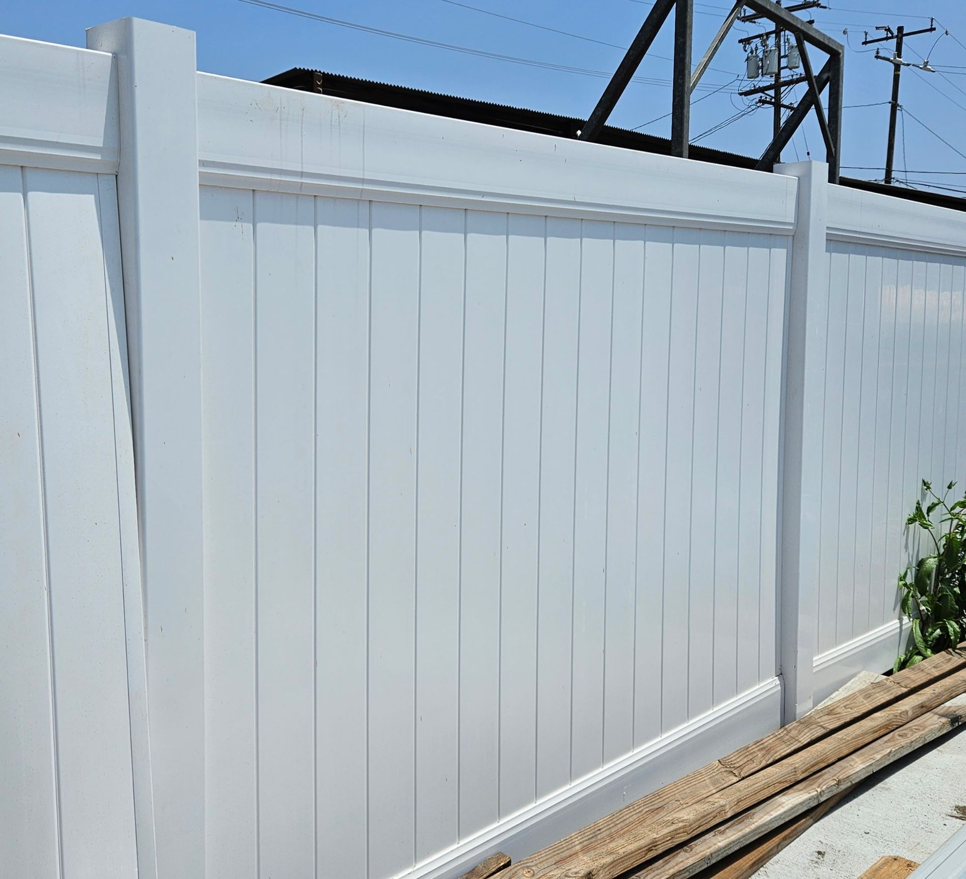 A white vinyl privacy fence stands outdoors under a clear blue sky, with a stack of wooden boards at its base.