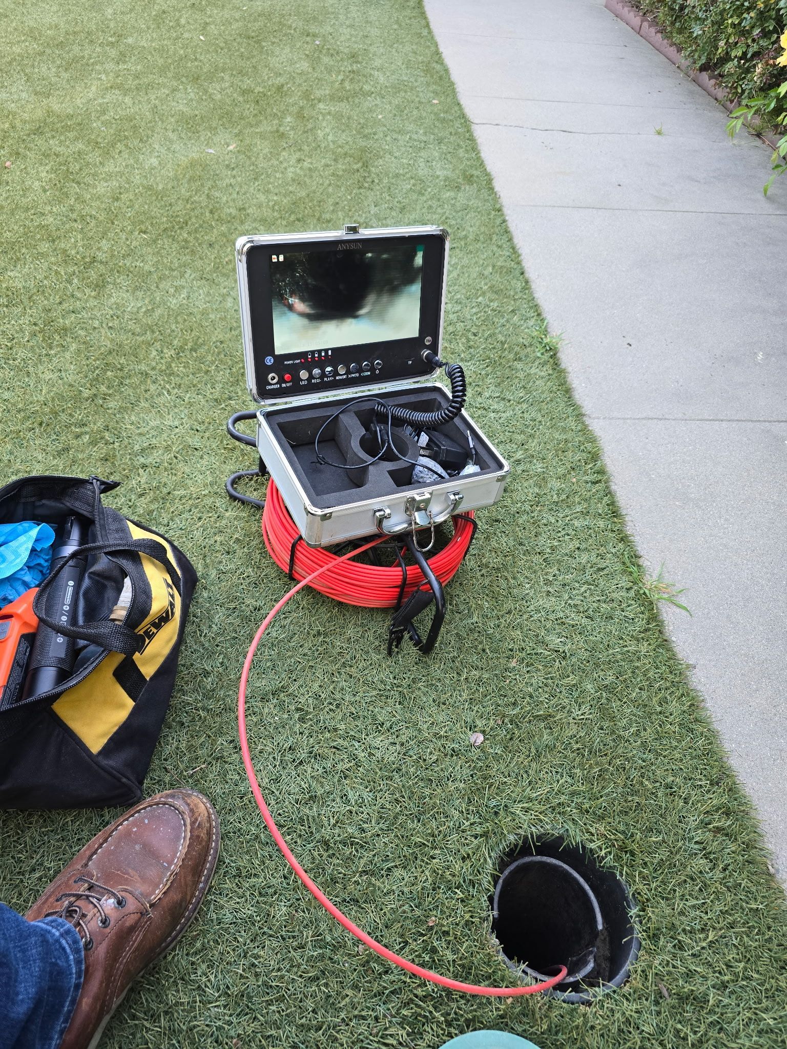 A sewer inspection camera on a lawn, connected to a pipe opening in the ground, with a tool bag and shoe nearby.