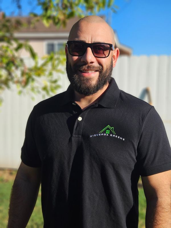 A smiling person with a beard and sunglasses wearing a black polo shirt with a green house logo, standing outdoors.