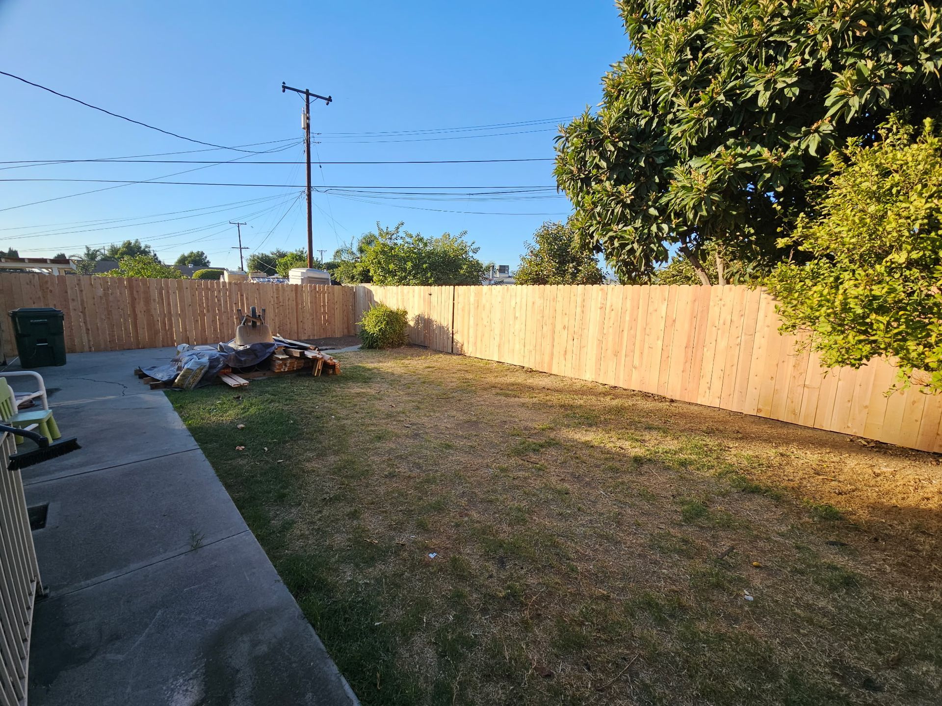 A sunny backyard with a concrete patio, a patch of dry grass, a new wooden fence, and a pile of debris in the corner.