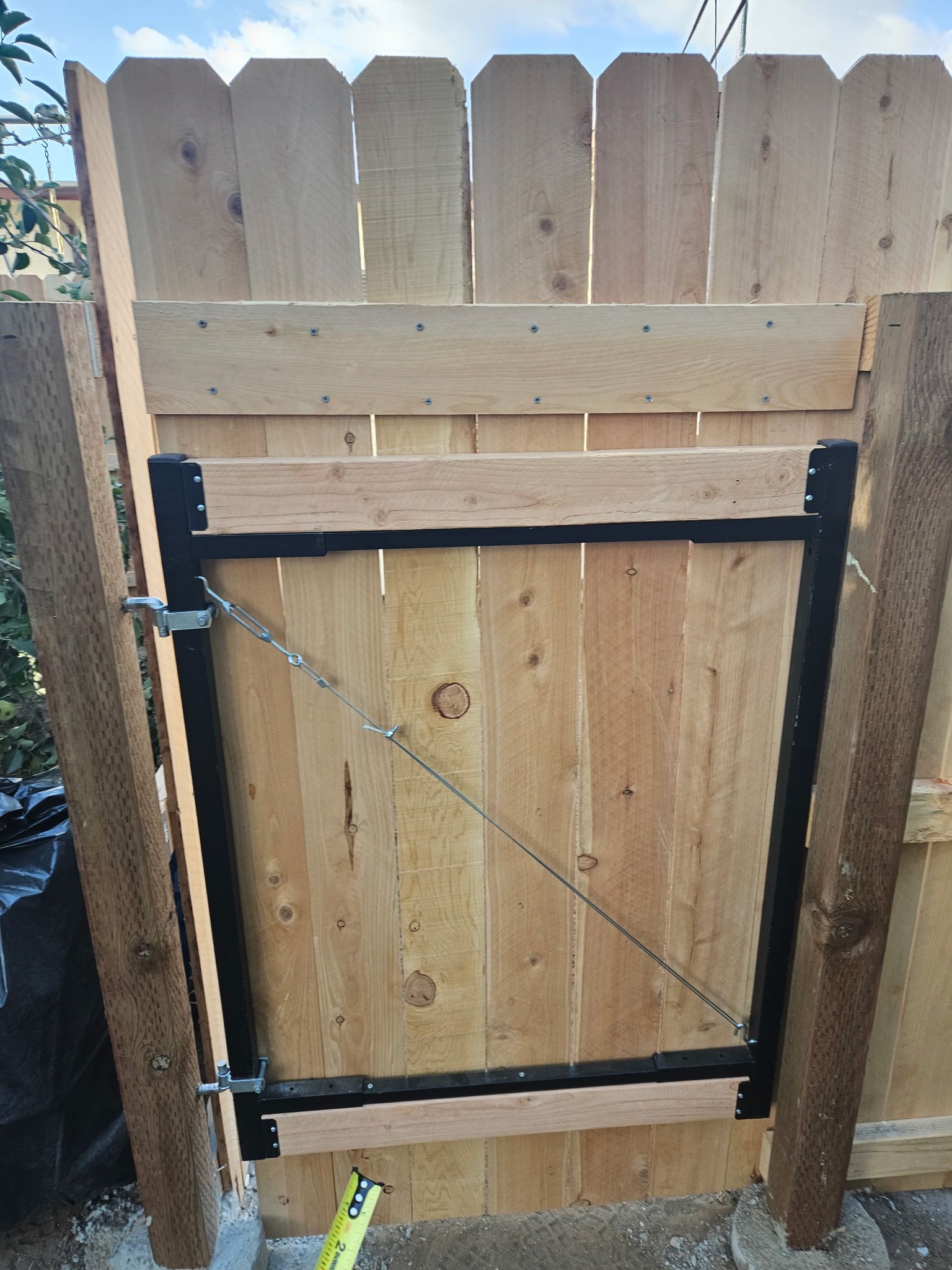 A wooden gate set within a fence, featuring a black metal frame and a diagonal tension cable.