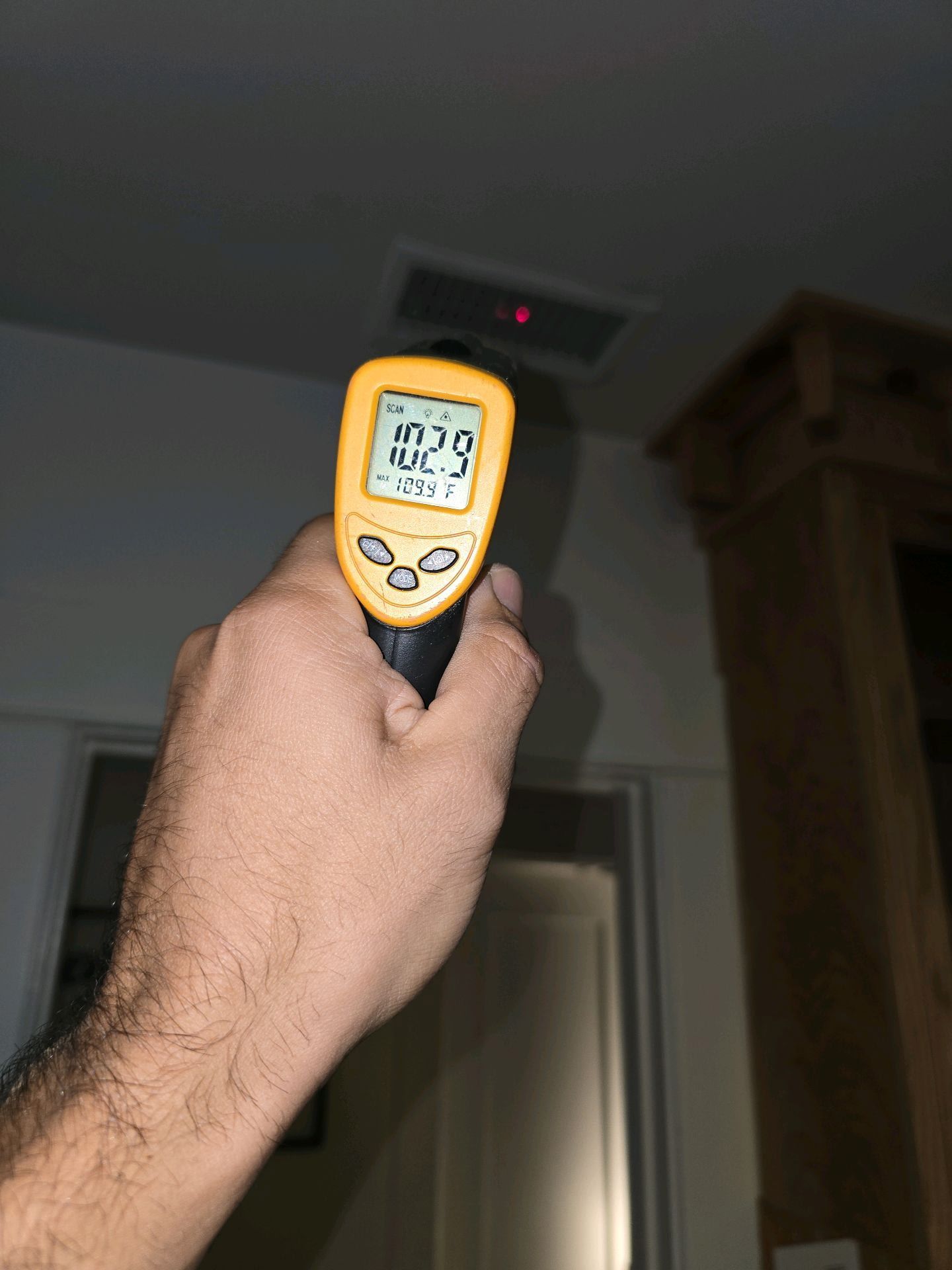 A hand holds a yellow infrared thermometer pointing at a ceiling vent, showing a reading of 102.9 degrees.