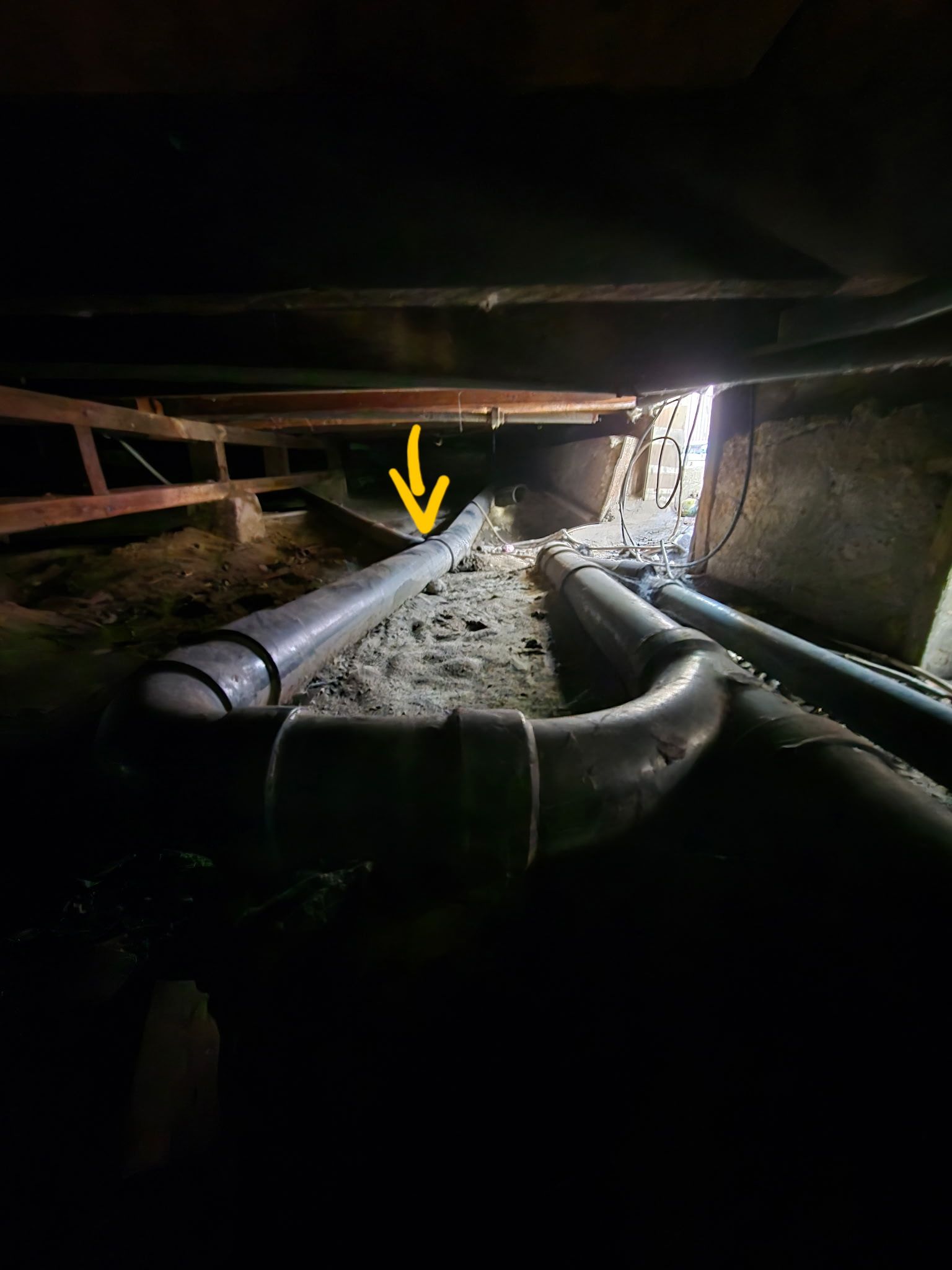 Crawl space with dark pipes, a yellow arrow pointing to a pipe, and debris-covered ground under a wooden structure.