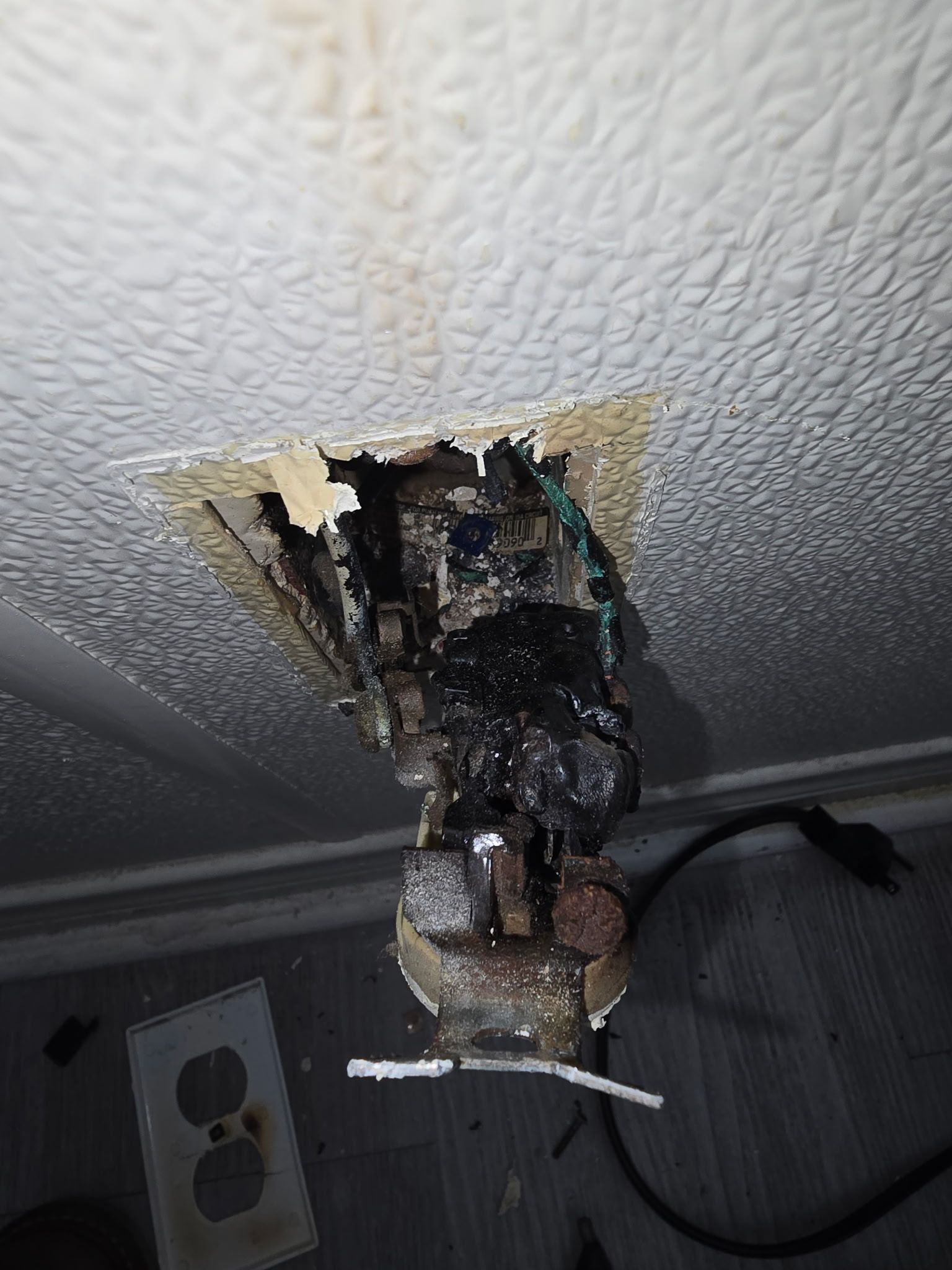 A wall outlet with severe fire damage, charred wiring, and melted plastic, with the cover plate lying on the floor.