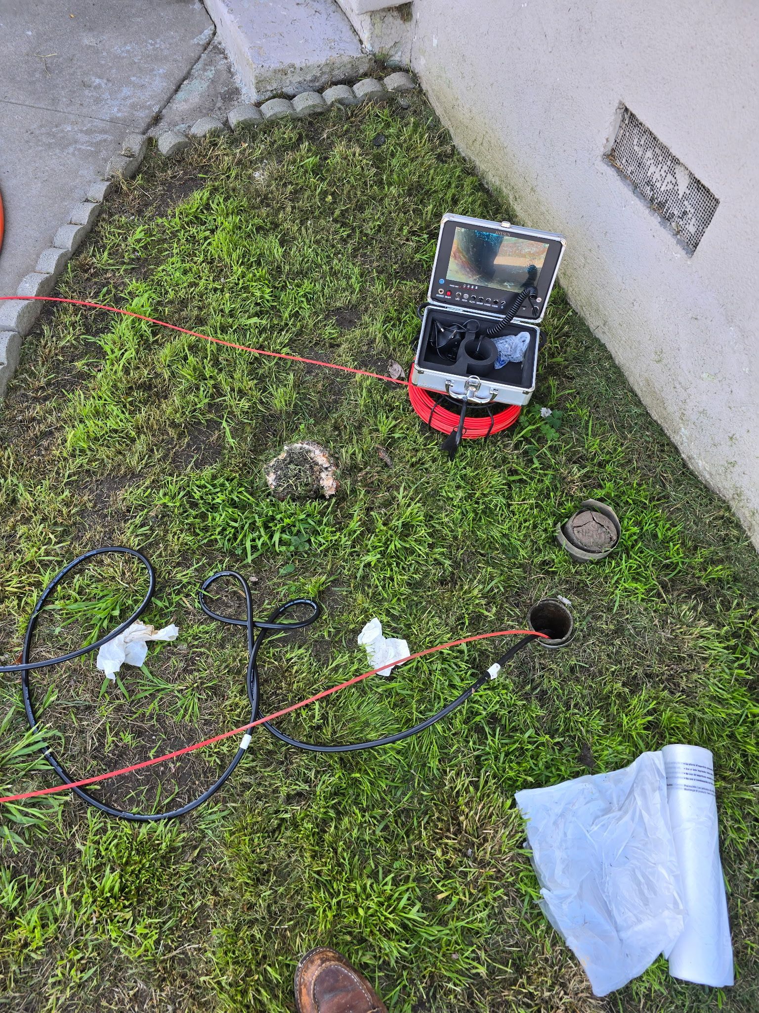 A sewer inspection camera system sits on a grassy patch next to a building wall, with a coiled cable and paper nearby.