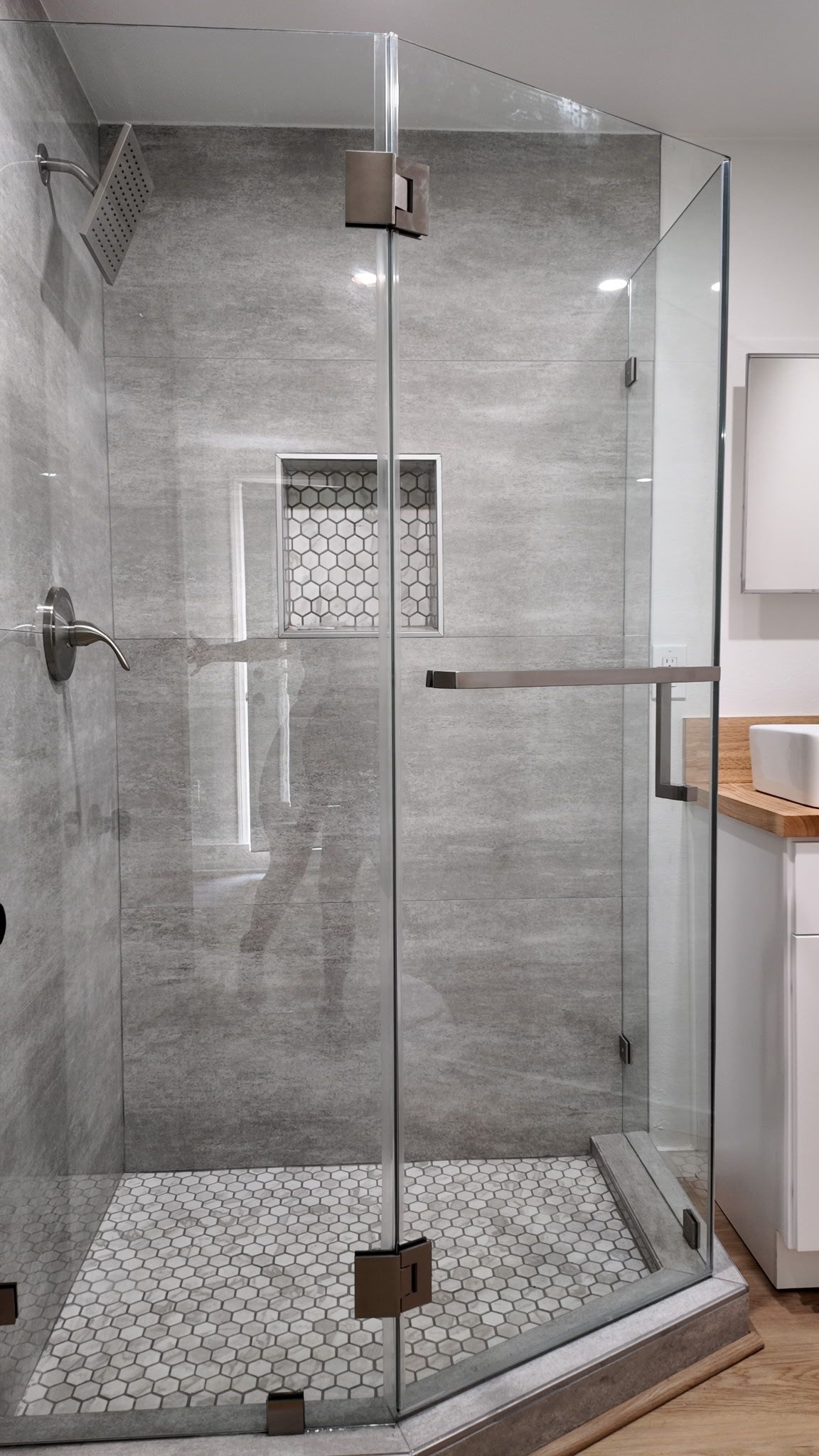 A modern glass shower enclosure with gray stone-look walls and a mosaic tile floor in a bright bathroom.