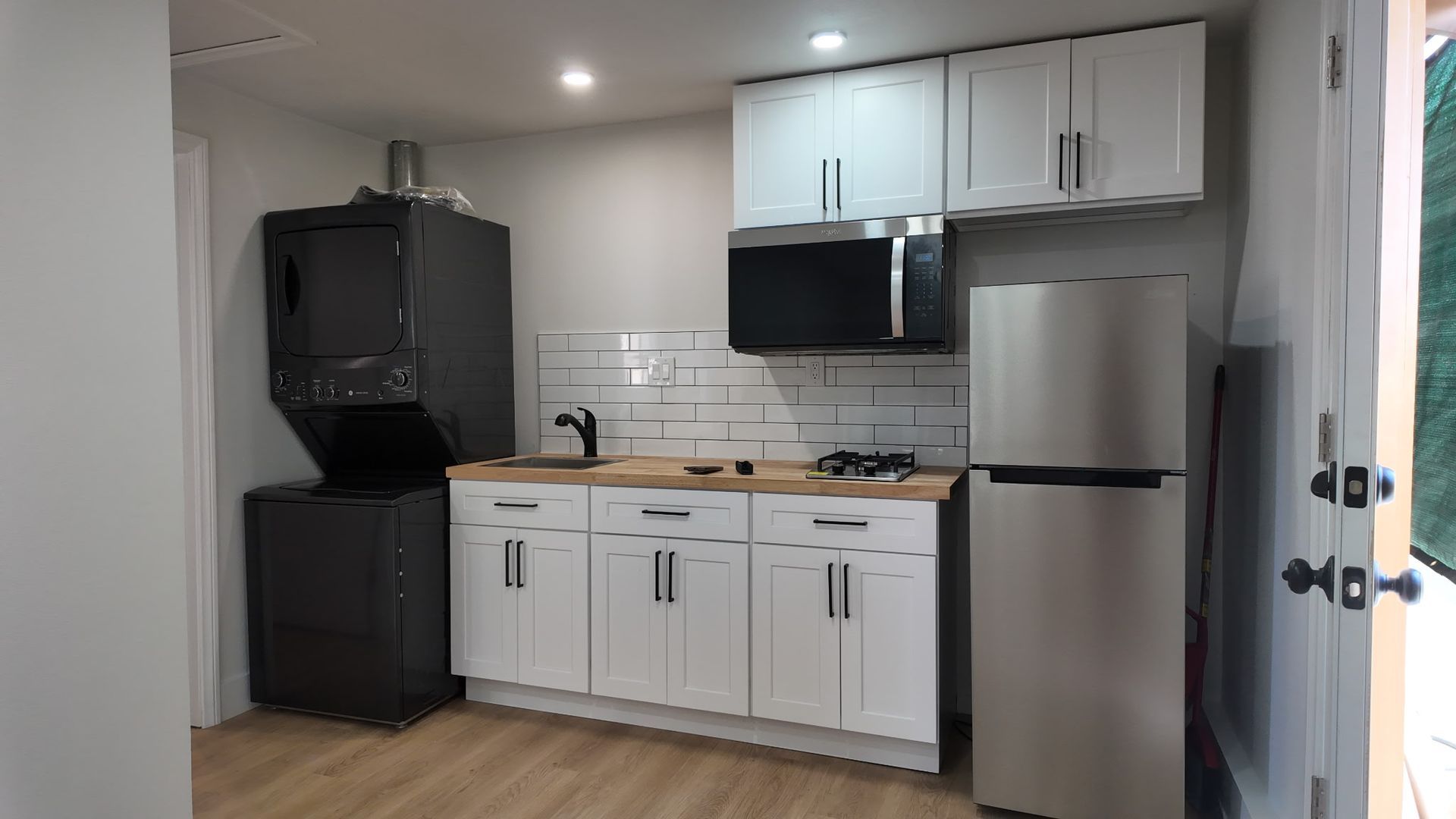 A modern kitchen features white cabinets, a stainless steel refrigerator, microwave, sink, and a dark stacked washer-dryer.