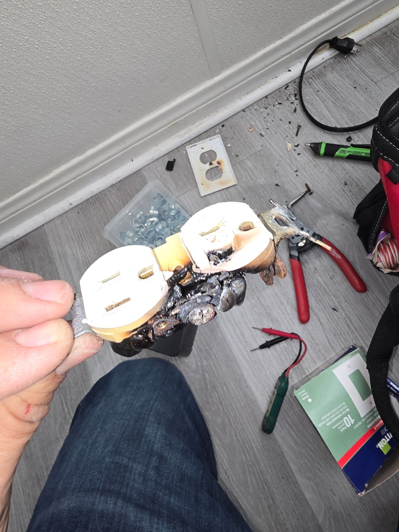 A hand holds a severely burned, damaged electrical outlet near a floor with scattered tools and a new outlet cover plate.