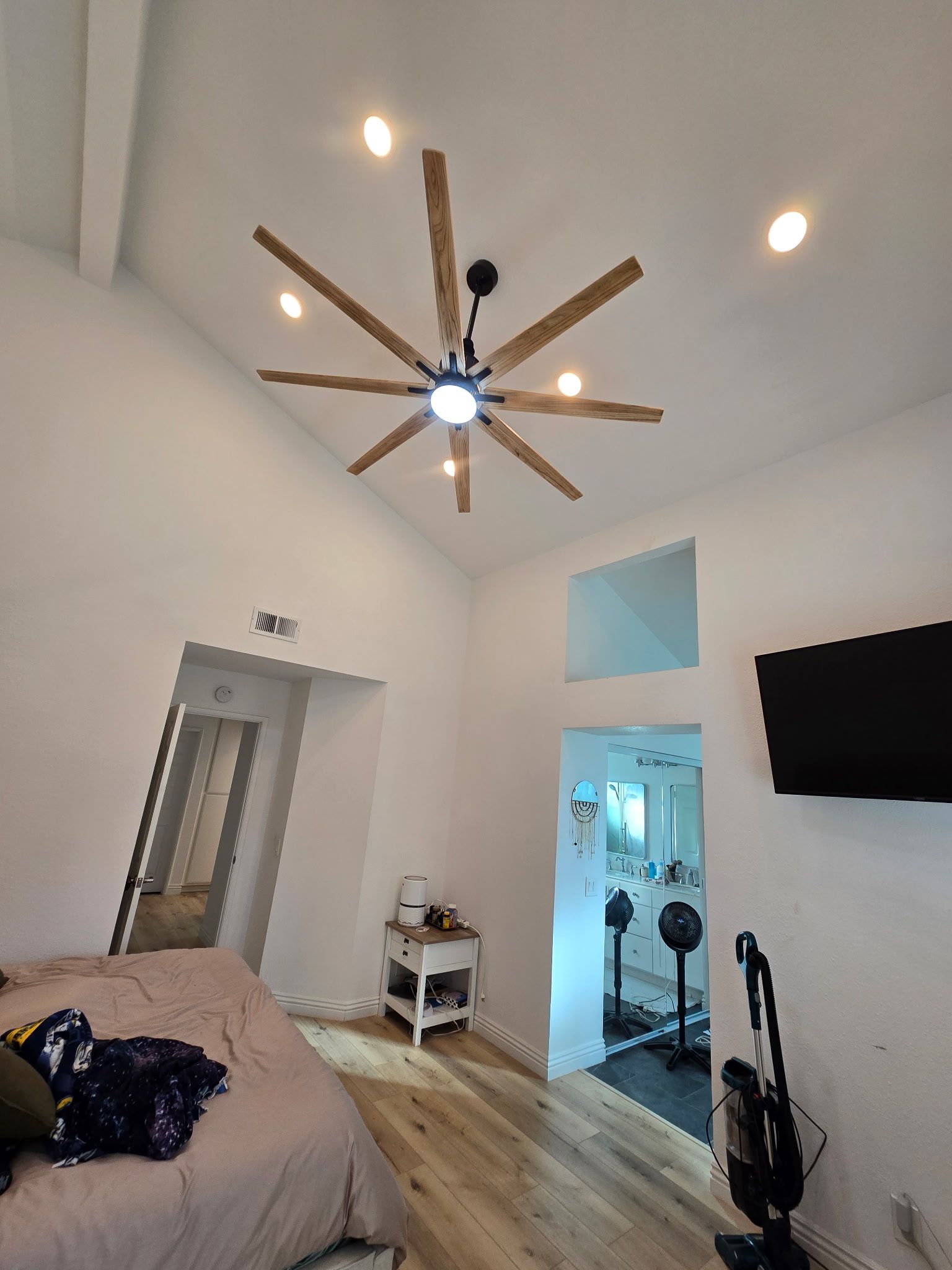 A bedroom interior featuring a large wooden ceiling fan, light-colored walls, wood floors, and a doorway to a bathroom.