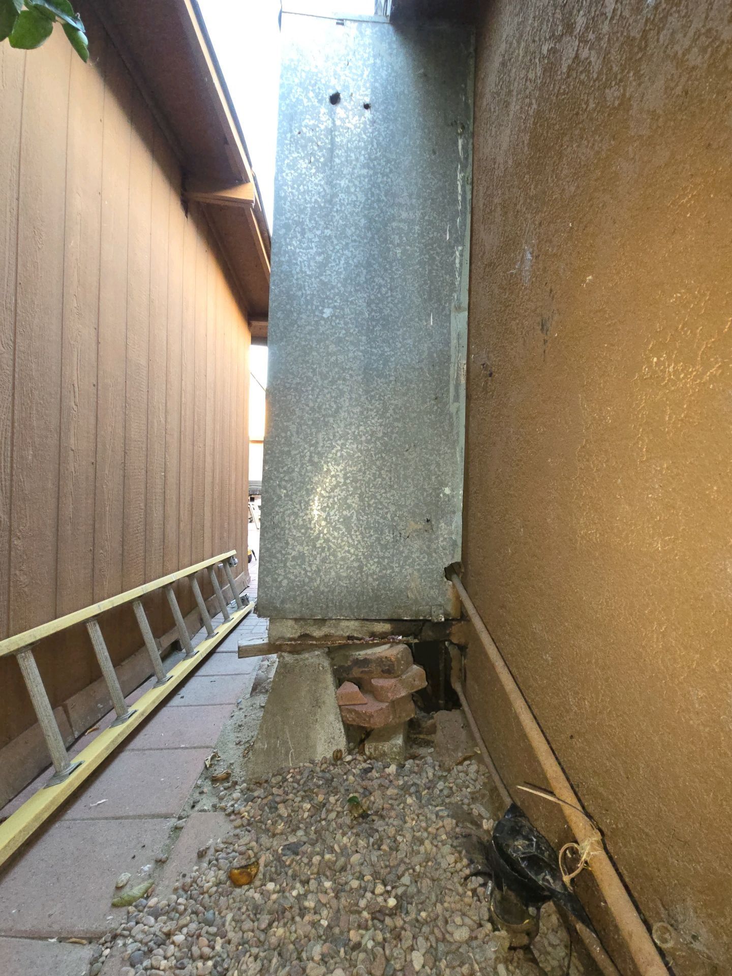 A narrow exterior walkway showing a metal duct chase resting on makeshift brick and stone supports against a house wall.