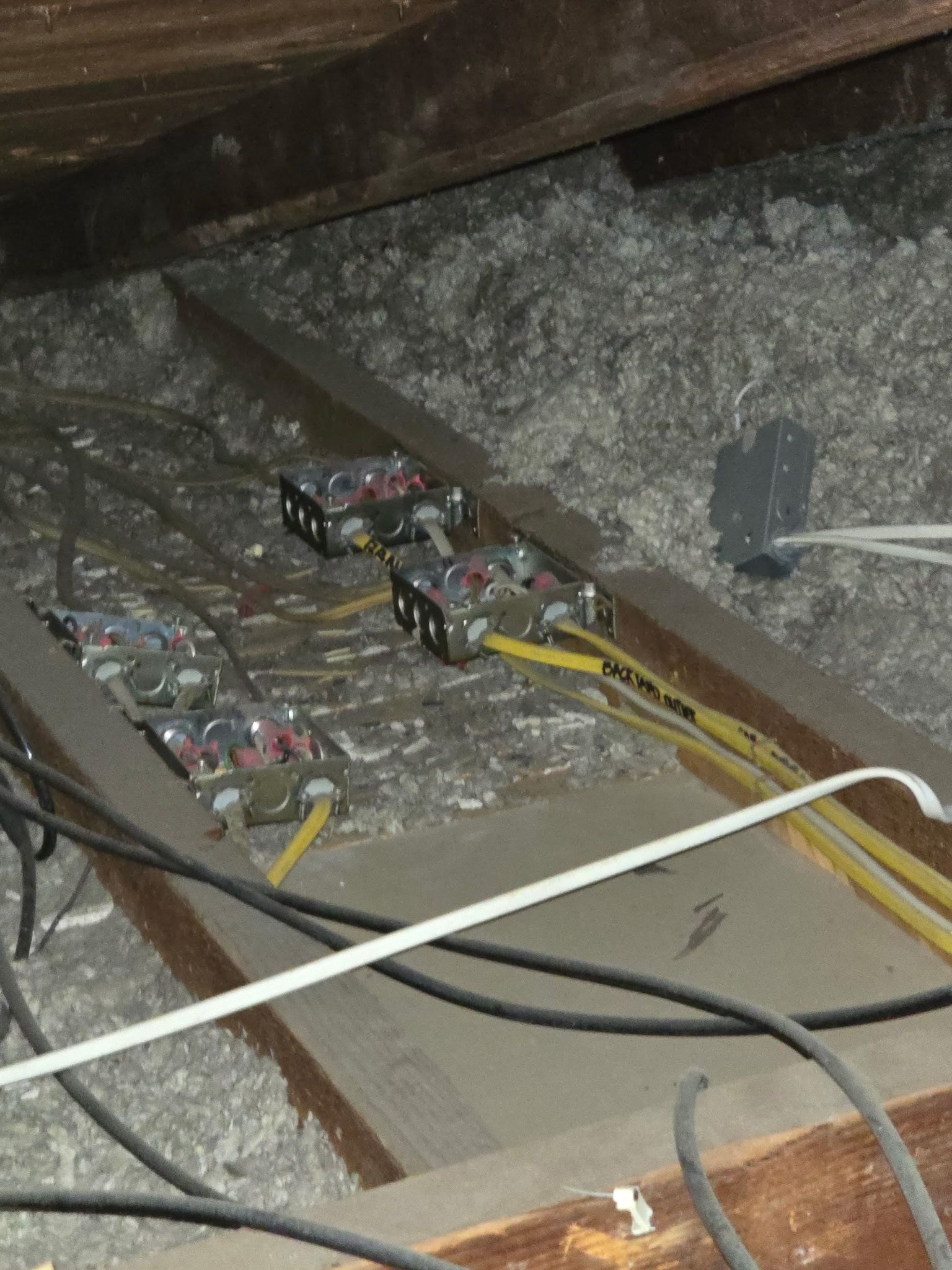 Electrical junction boxes with connected wires resting on wooden joists amidst loose-fill insulation in an attic.
