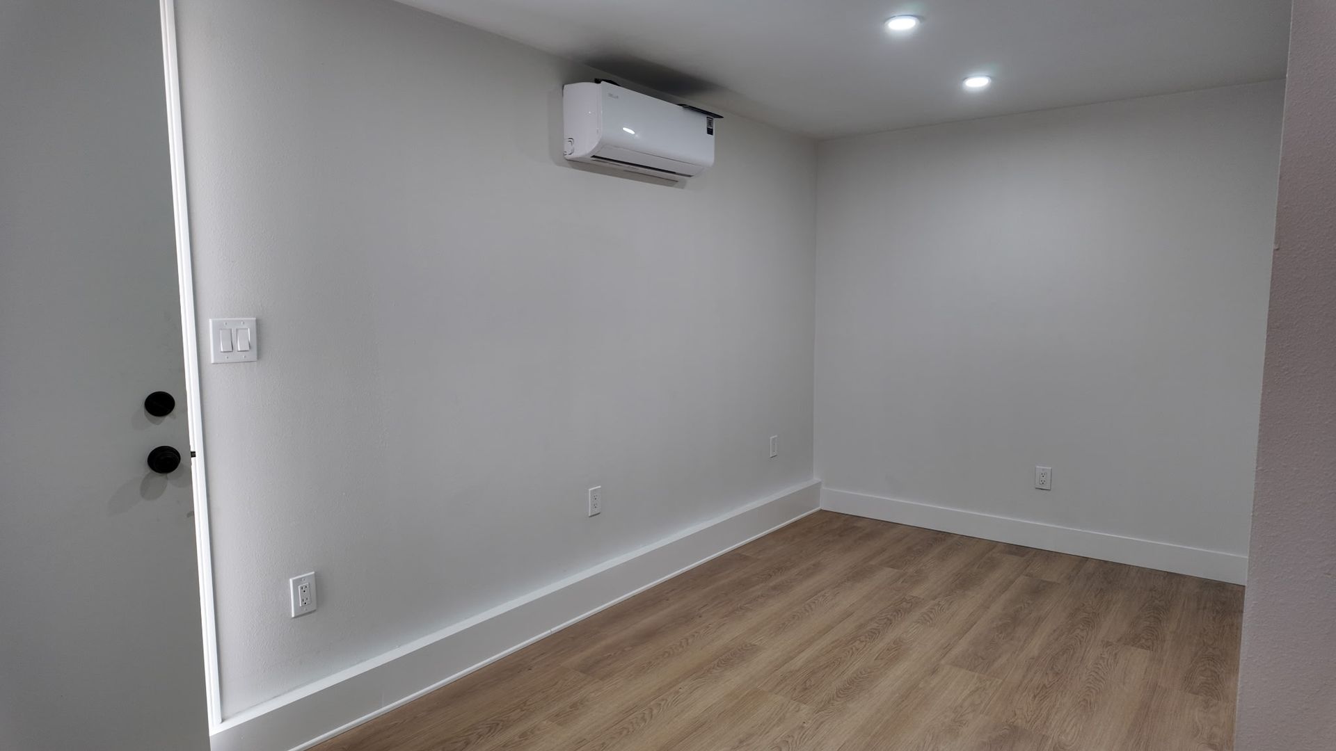 A bright, empty room with light wood-look flooring, white walls, a mounted air conditioner, and two recessed lights.