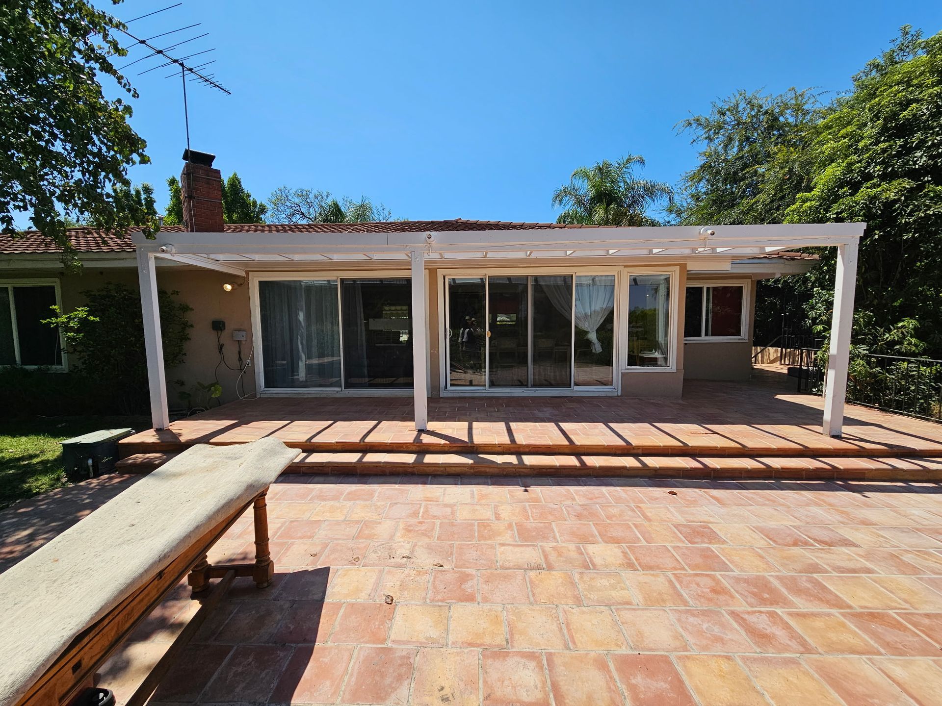 A house with a large terracotta-tiled patio, a pergola, and sliding glass doors under a clear blue sky.