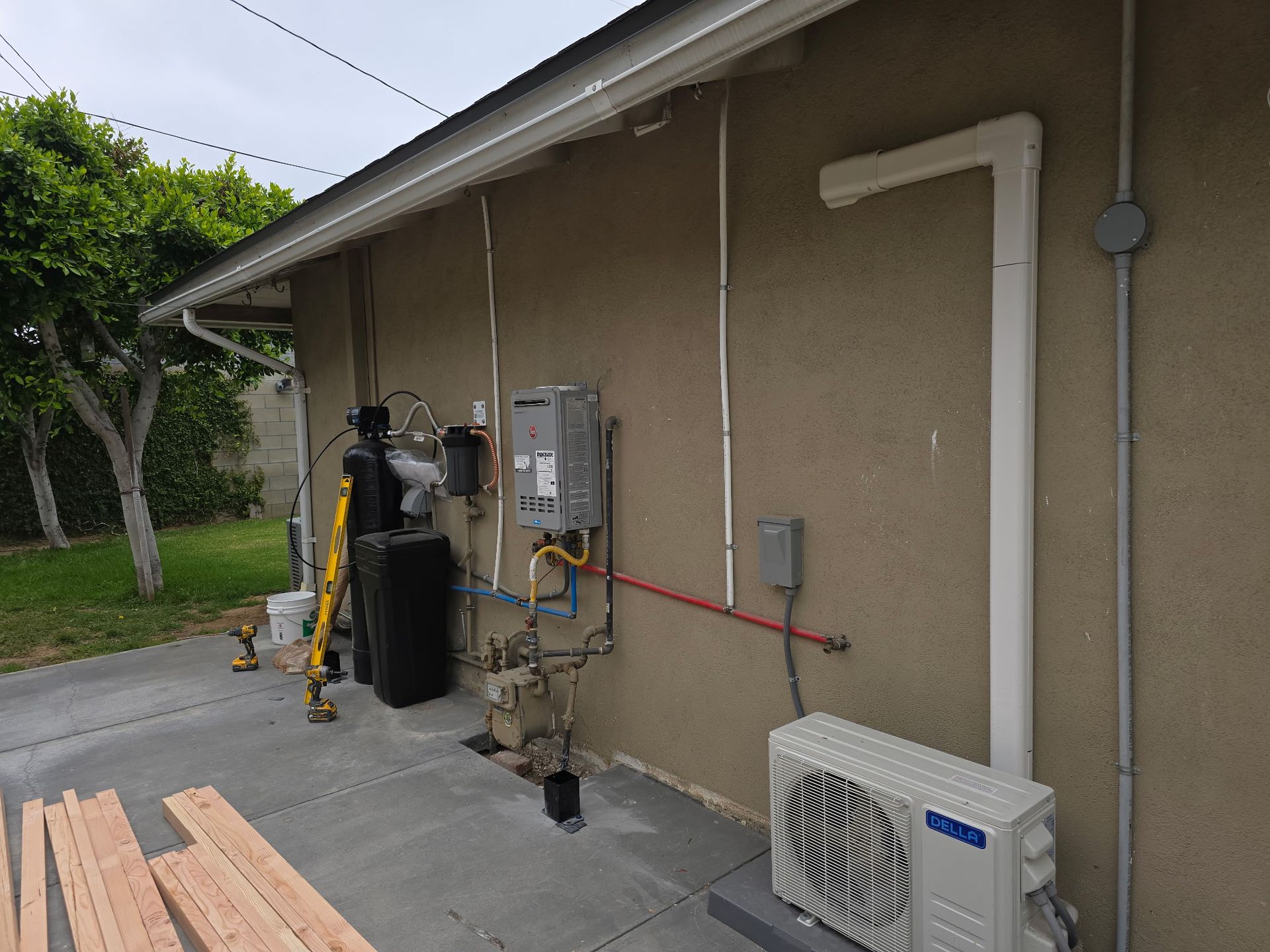 Exterior wall with a tankless water heater, water filtration system, and an outdoor HVAC unit on a concrete patio.