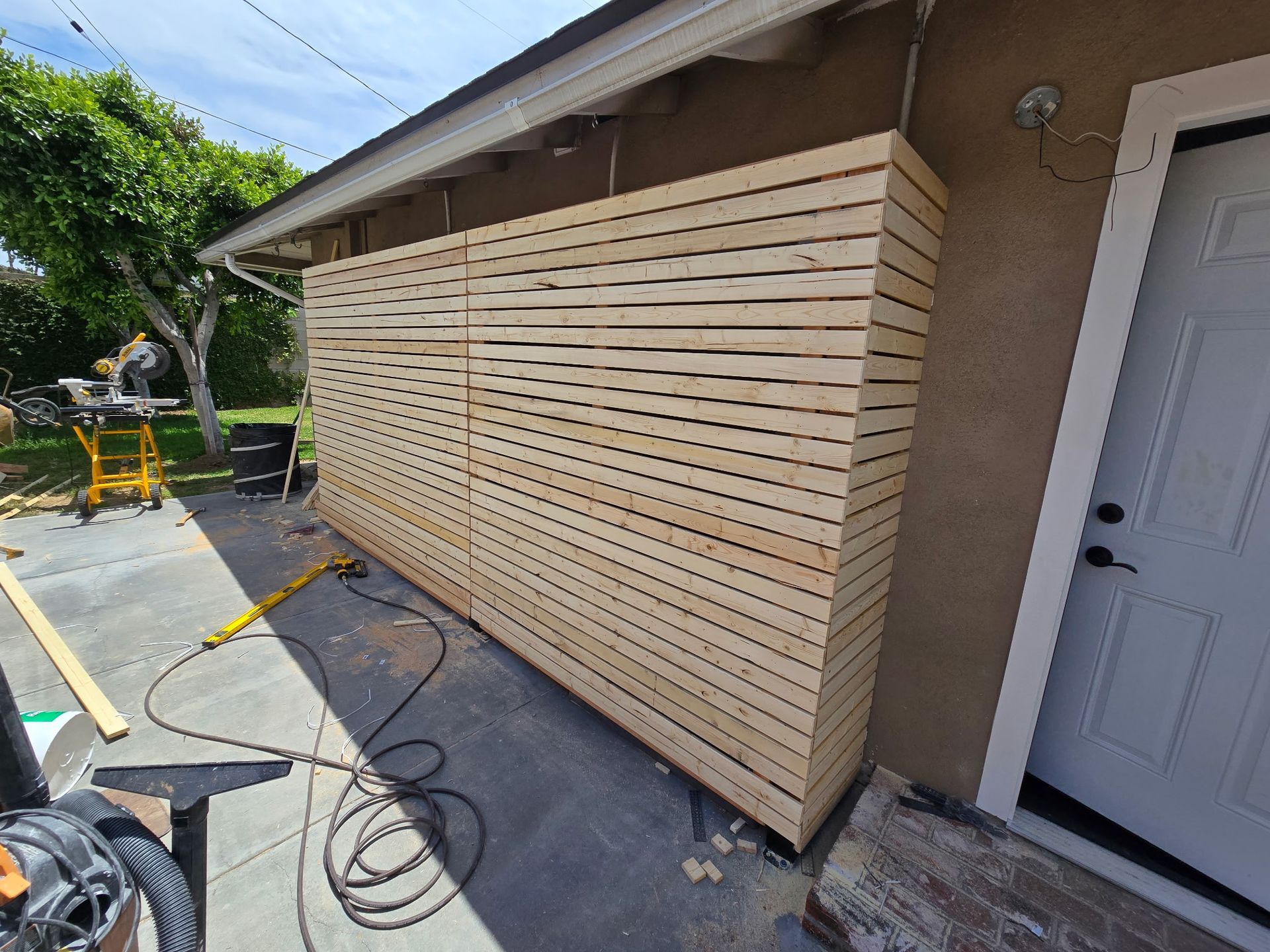 A horizontal wood slat privacy screen wall installed along the exterior wall of a house near a doorway on a patio.