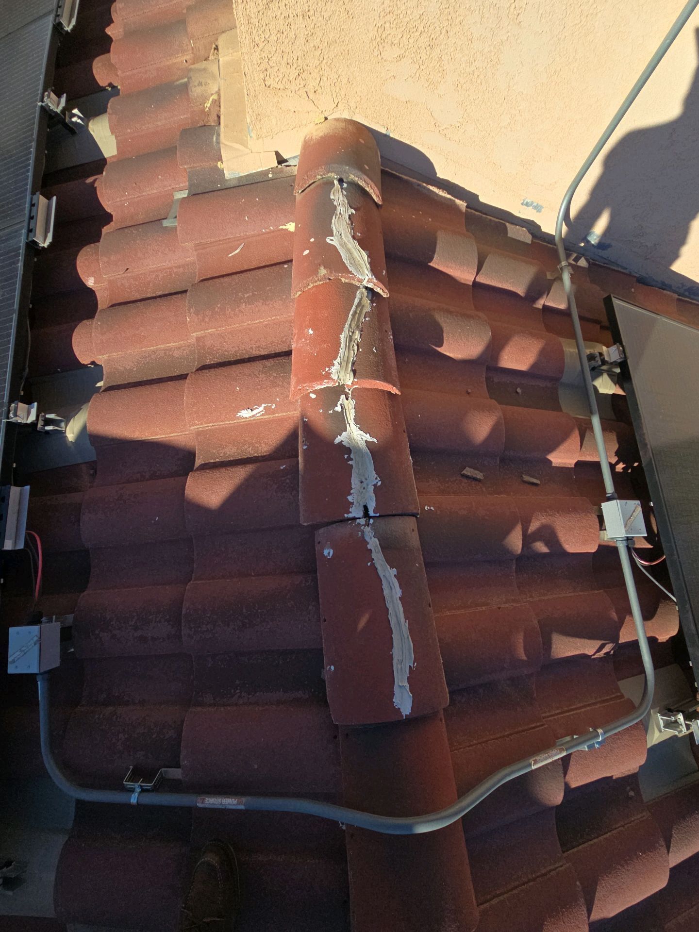 A metal conduit runs across red clay roof tiles toward a solar panel installation, with sealant applied to the roof ridge.