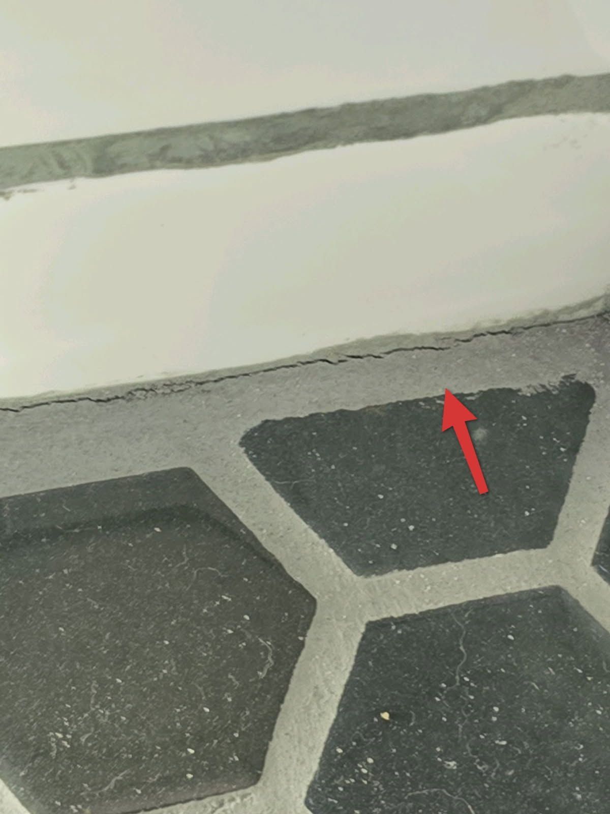 A close-up of a crack where a white wall meets dark gray hexagonal floor tiles, pointed out by a red arrow.