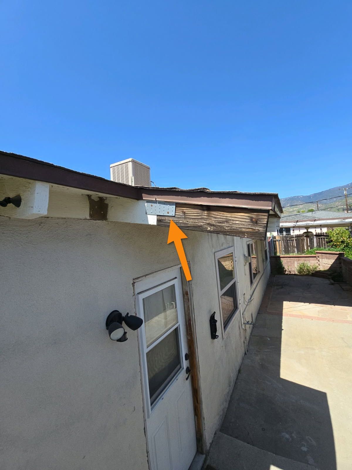 An orange arrow points to exposed wooden framing and damaged roofing fascia on the side of a stucco building.