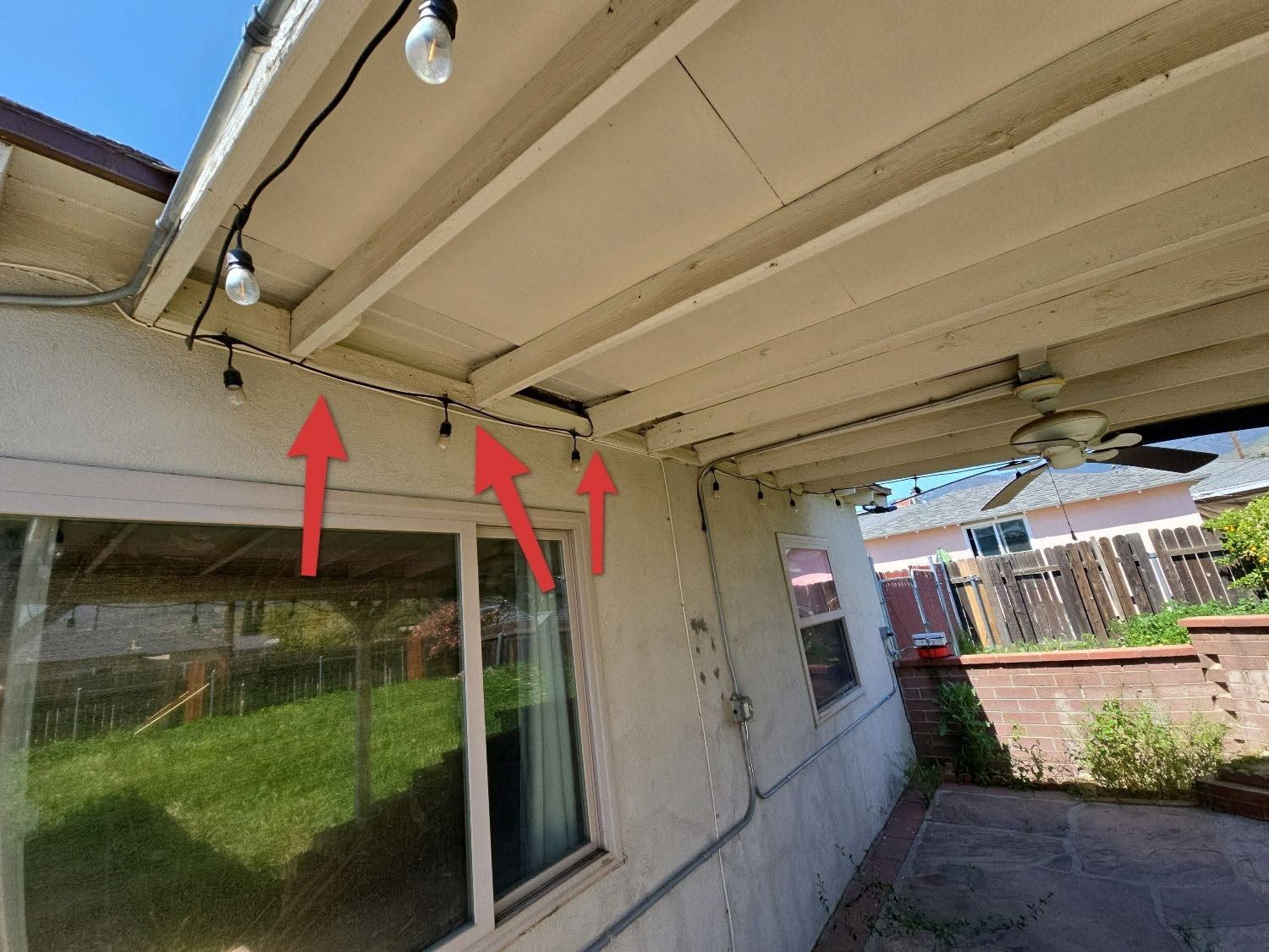 Patio area showing three red arrows pointing to string lights mounted along the ceiling beams of a house overhang.