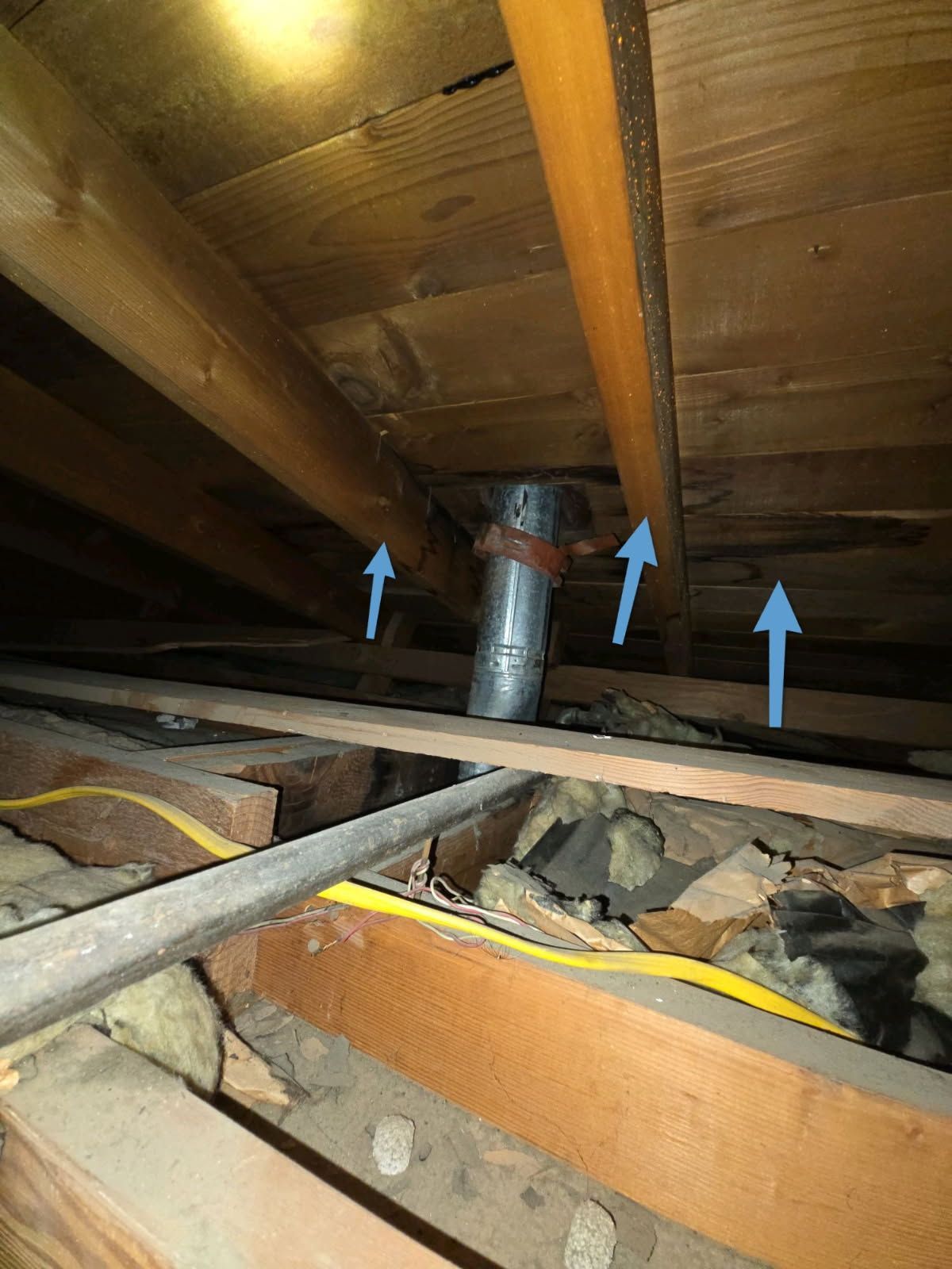Attic view of a metal vent pipe passing through the roof rafters, with blue arrows pointing to gaps in the roof decking.