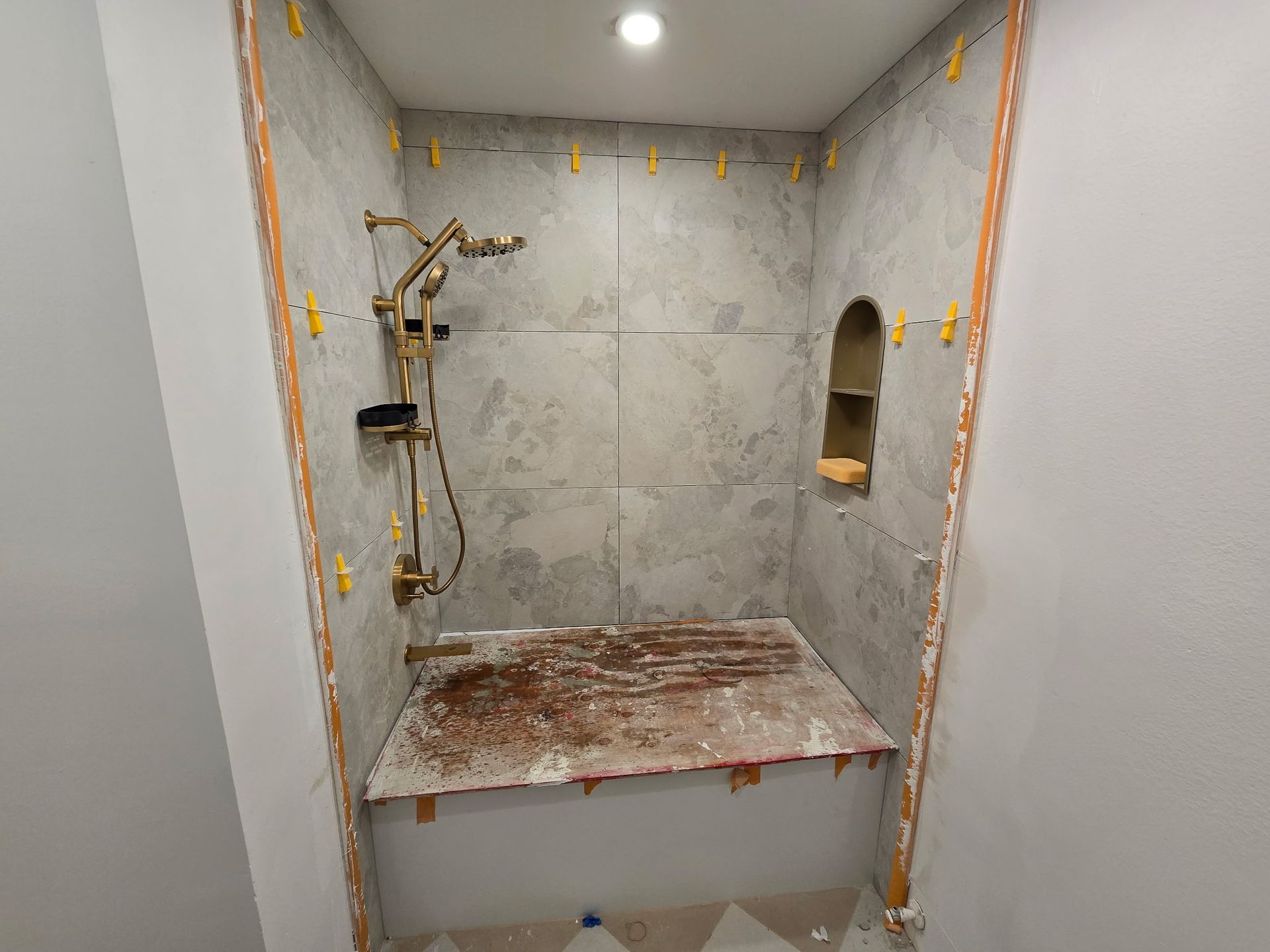 Small unfinished shower alcove with gray tile, brass fixtures, and a rough ledge under a ceiling light