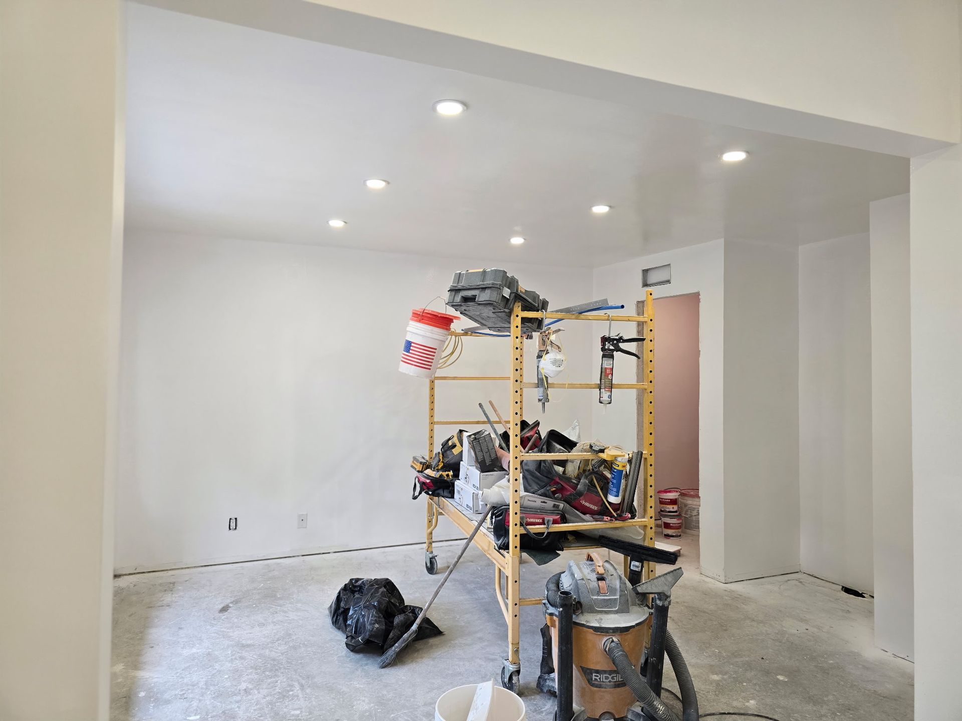Renovation room with white walls, ceiling lights, scaffolding, tools, and construction debris on the floor