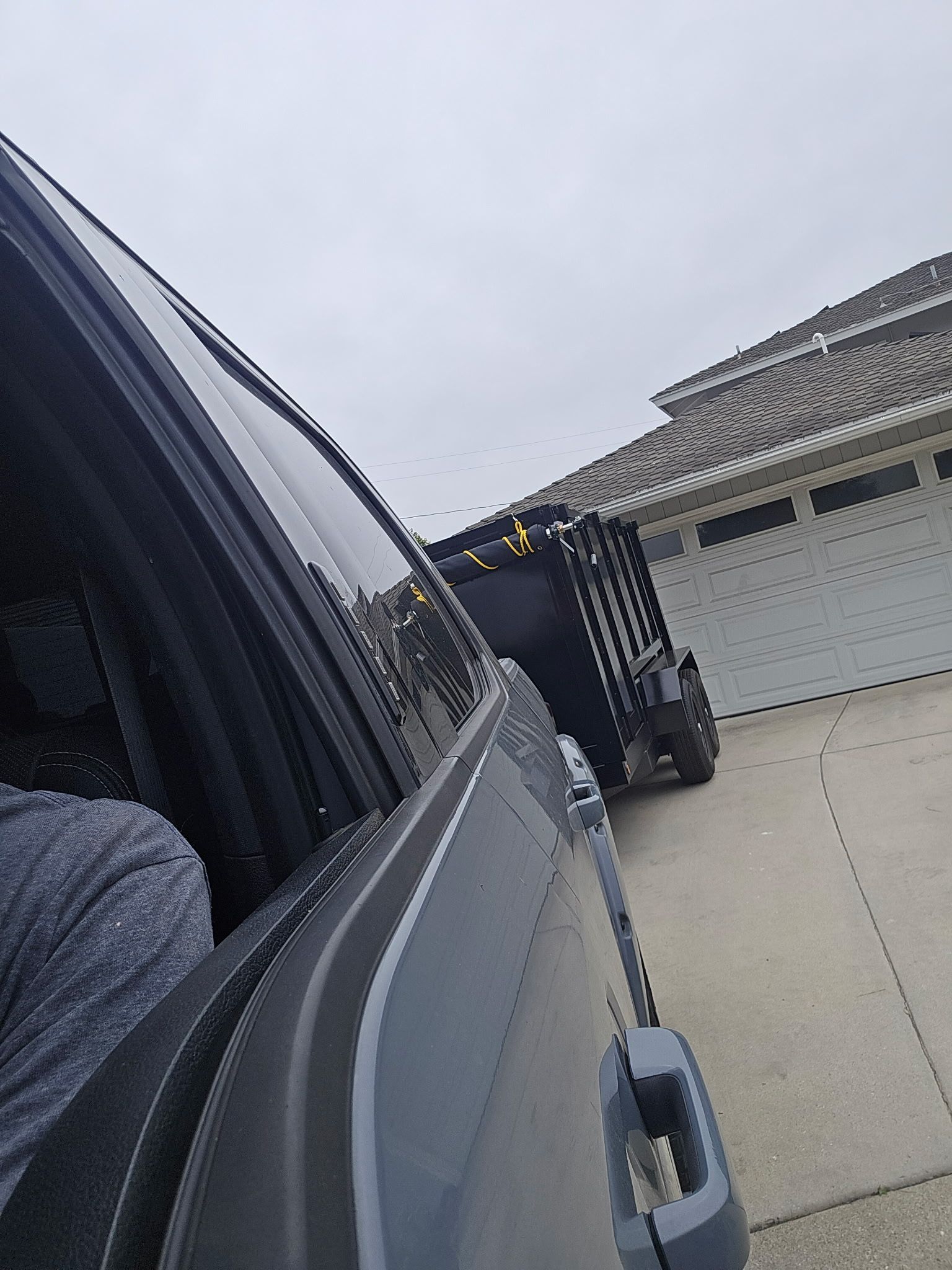 Car window view of a driveway with a black trailer and a house garage on an overcast day