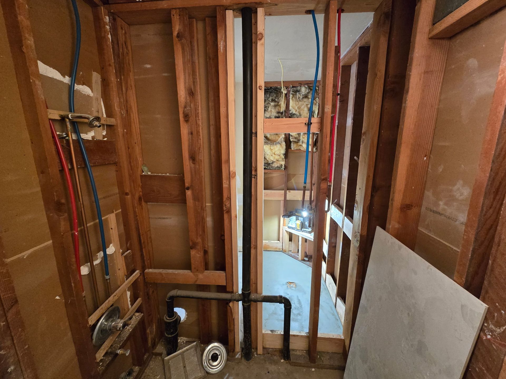 Exposed wall framing in a gutted room with pipes, wiring, and a doorway to a lit space beyond