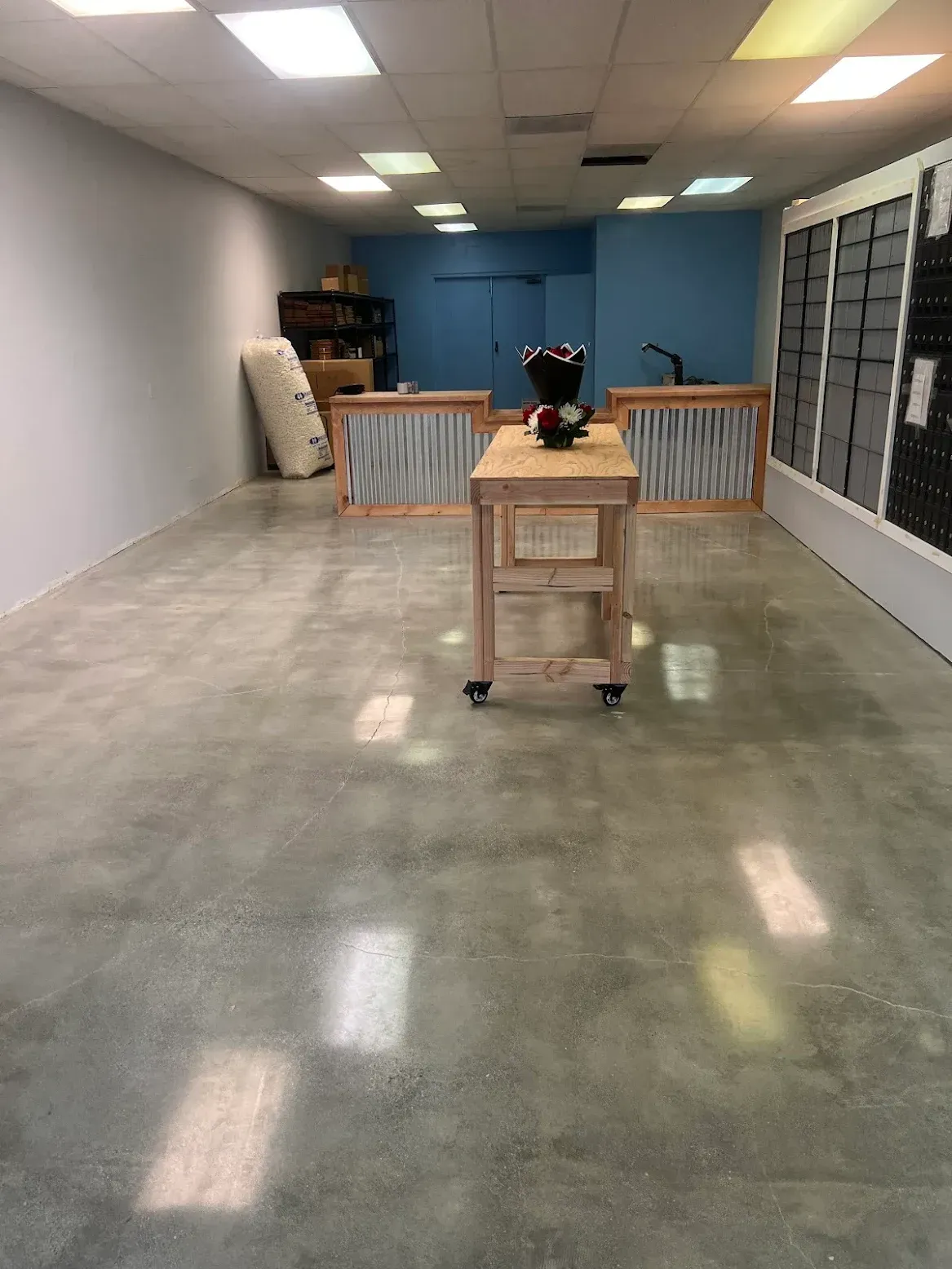 A bright, empty retail space with a polished concrete floor, a wooden central workbench, and rows of wall-mounted shelves.