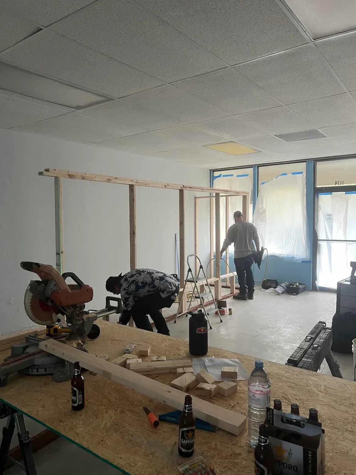 Two people working on a wooden frame inside a room with a miter saw and tools on a workbench in the foreground.