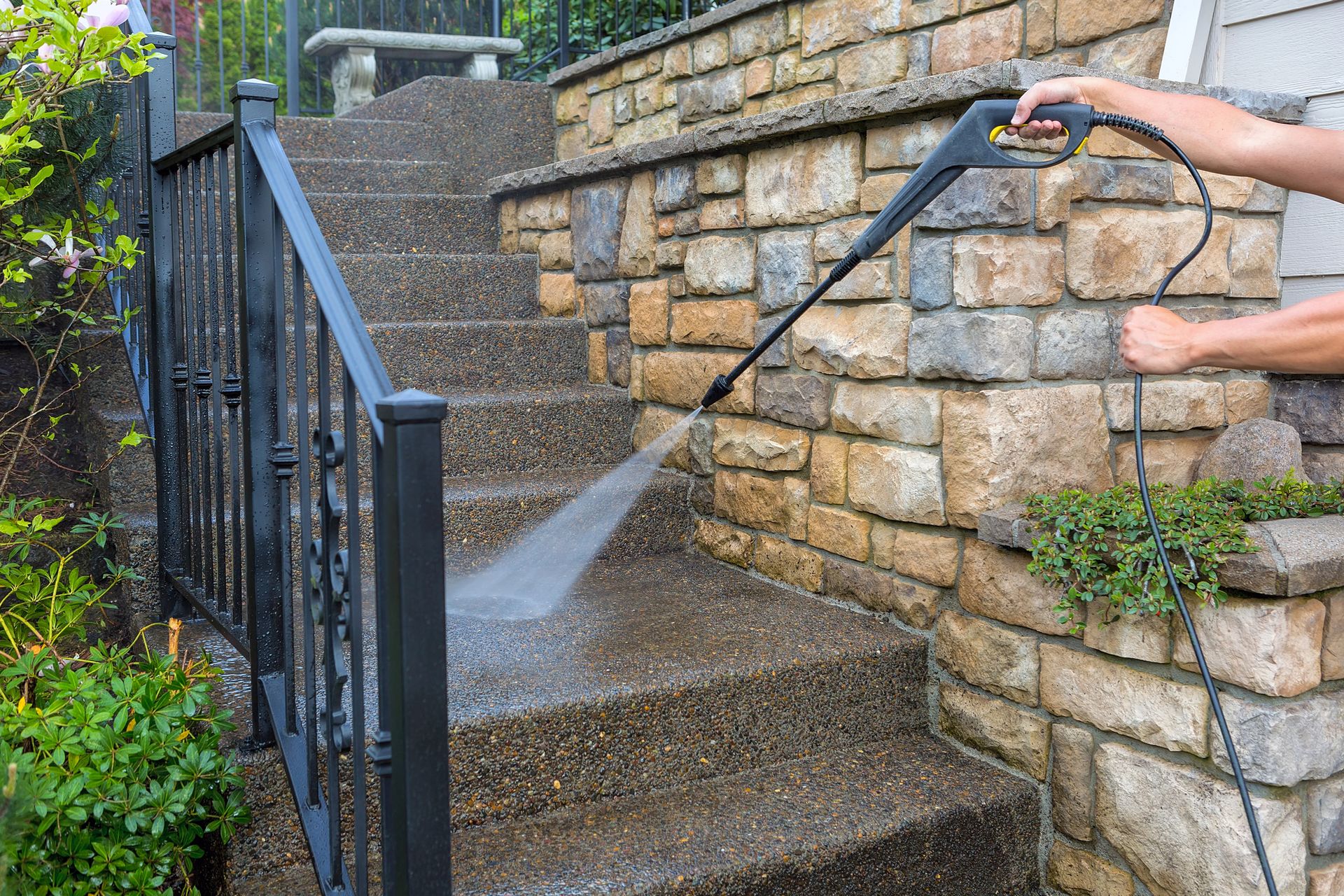 A person uses a pressure washer to clean stone steps next to a metal railing and a stone retaining wall.